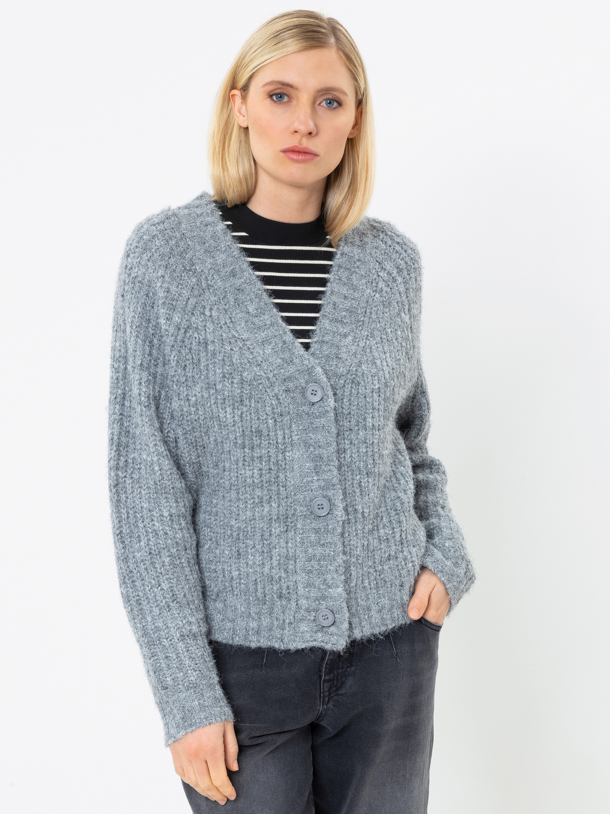 Eine Frau mit glattem blondem Haar trägt eine graue Strickjacke mit großen Knöpfen über einem schwarz-weiß gestreiften Shirt, dazu eine dunkle Jeans, und steht vor einem schlichten weißen Hintergrund.