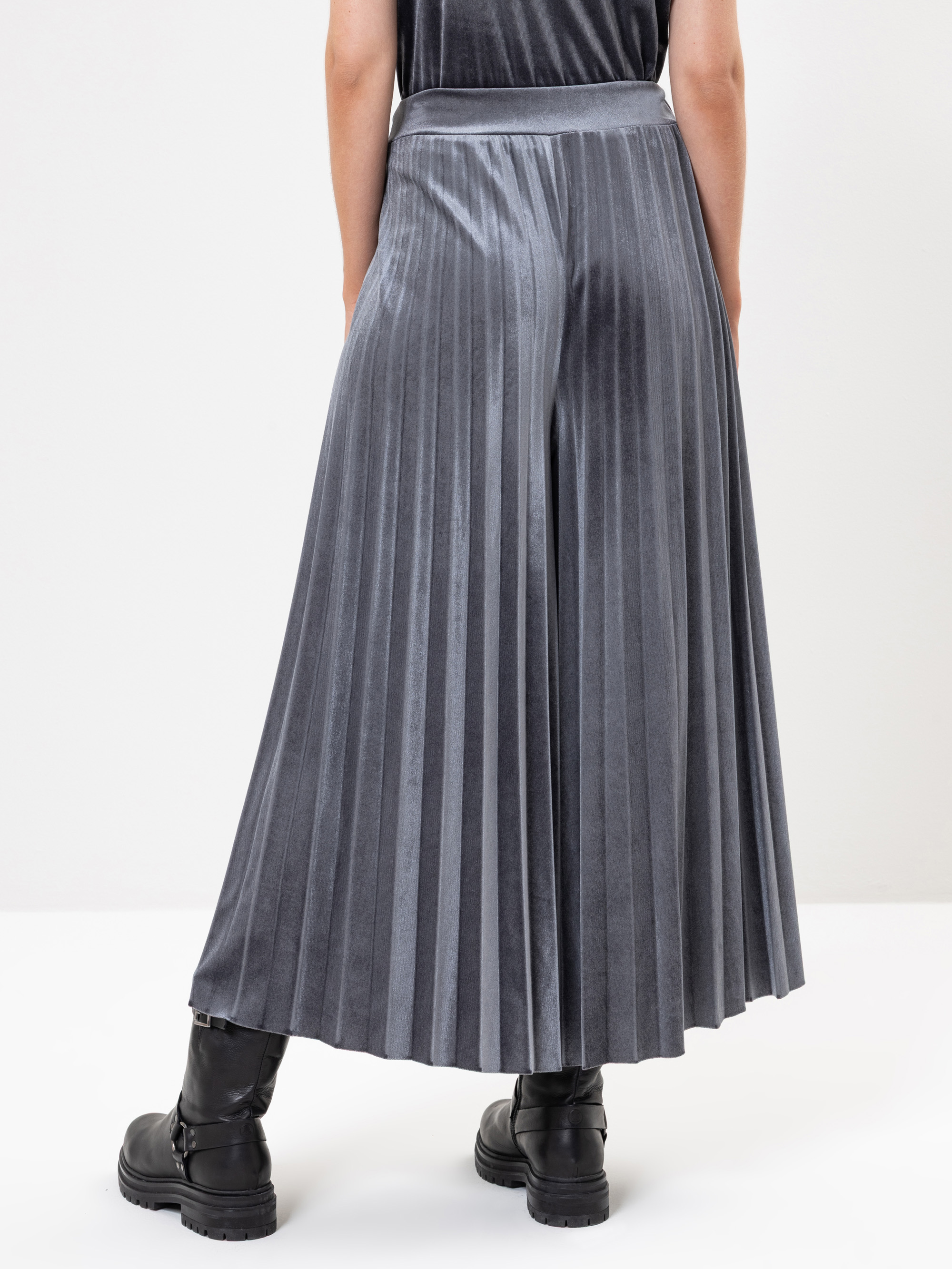 Eine Person, die einen langen, grauen Faltenrock und schwarze Stiefel trägt, steht mit dem Rücken zur Kamera vor einem schlichten weißen Hintergrund.