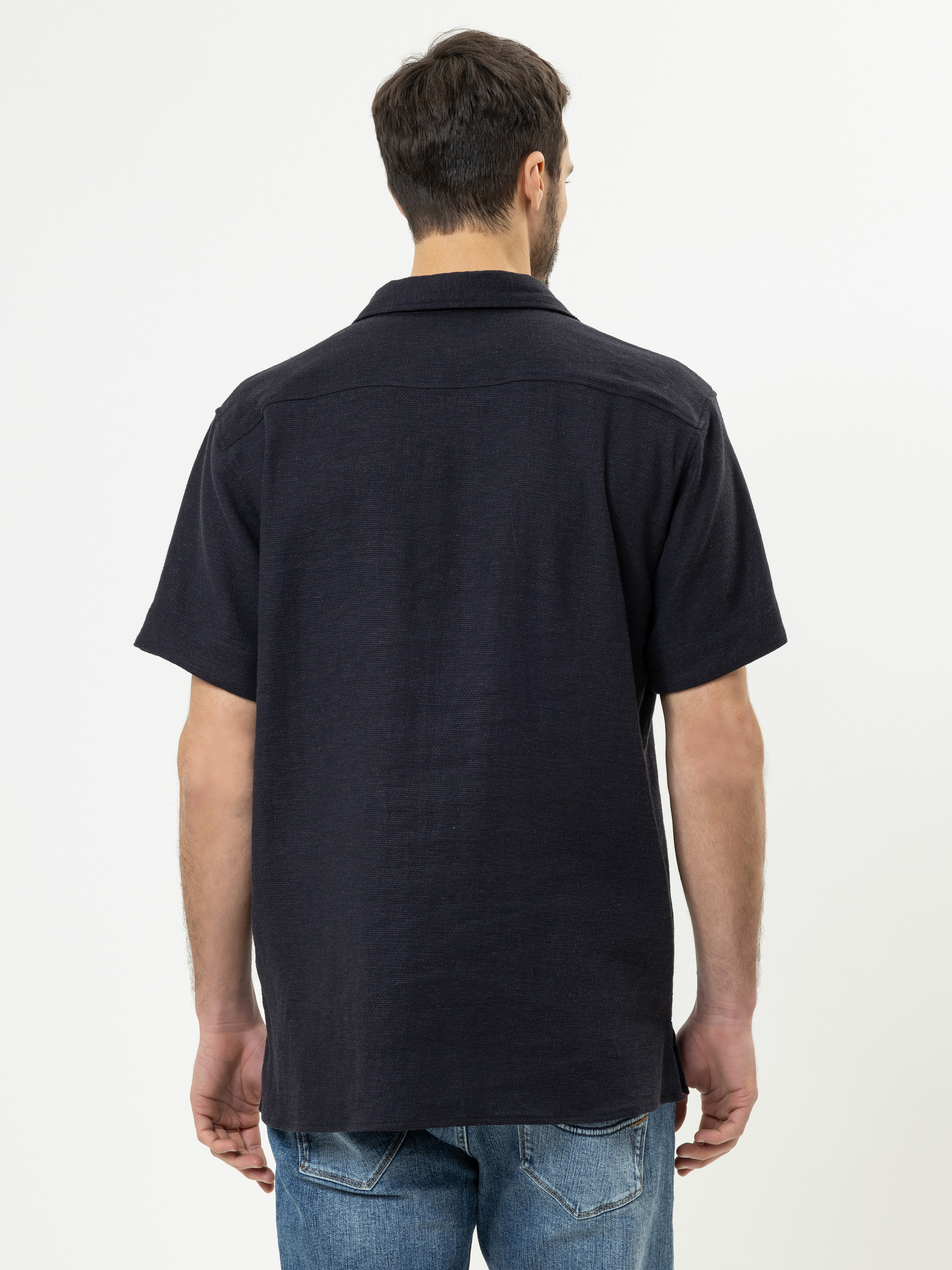 Ein Mann mit kurzen braunen Haaren steht und schaut von der Kamera weg. Er trägt ein kurzärmeliges, dunkles Hemd und eine blaue Jeans vor einem schlichten weißen Hintergrund.