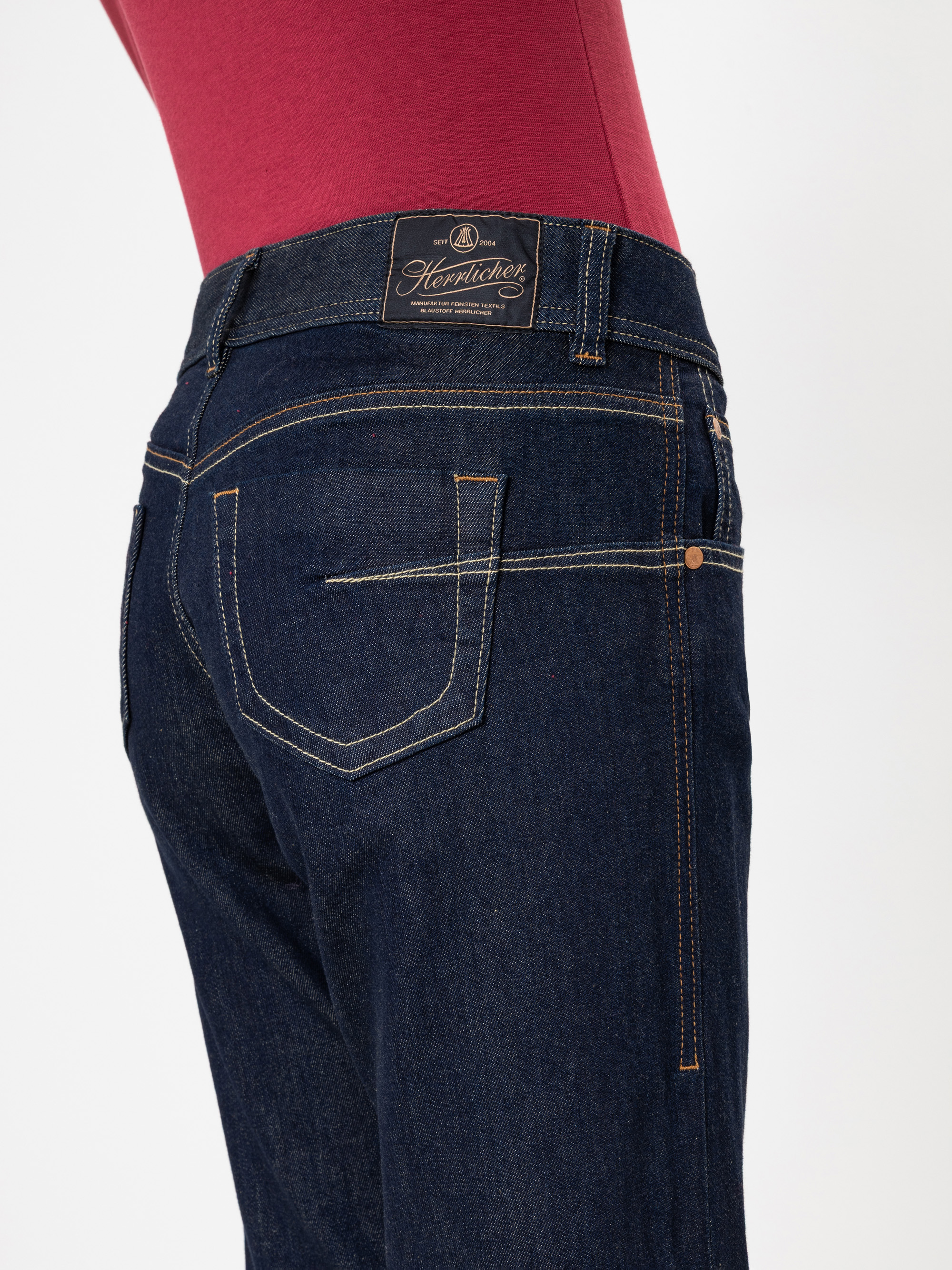 Eine Person trägt eine dunkelblaue Jeans und ein rotes Oberteil, von der Taille abwärts und von hinten und von der Seite gesehen. Die Jeans hat Kontrastnähte und einen braunen Markenaufnäher aus Leder am Bund.