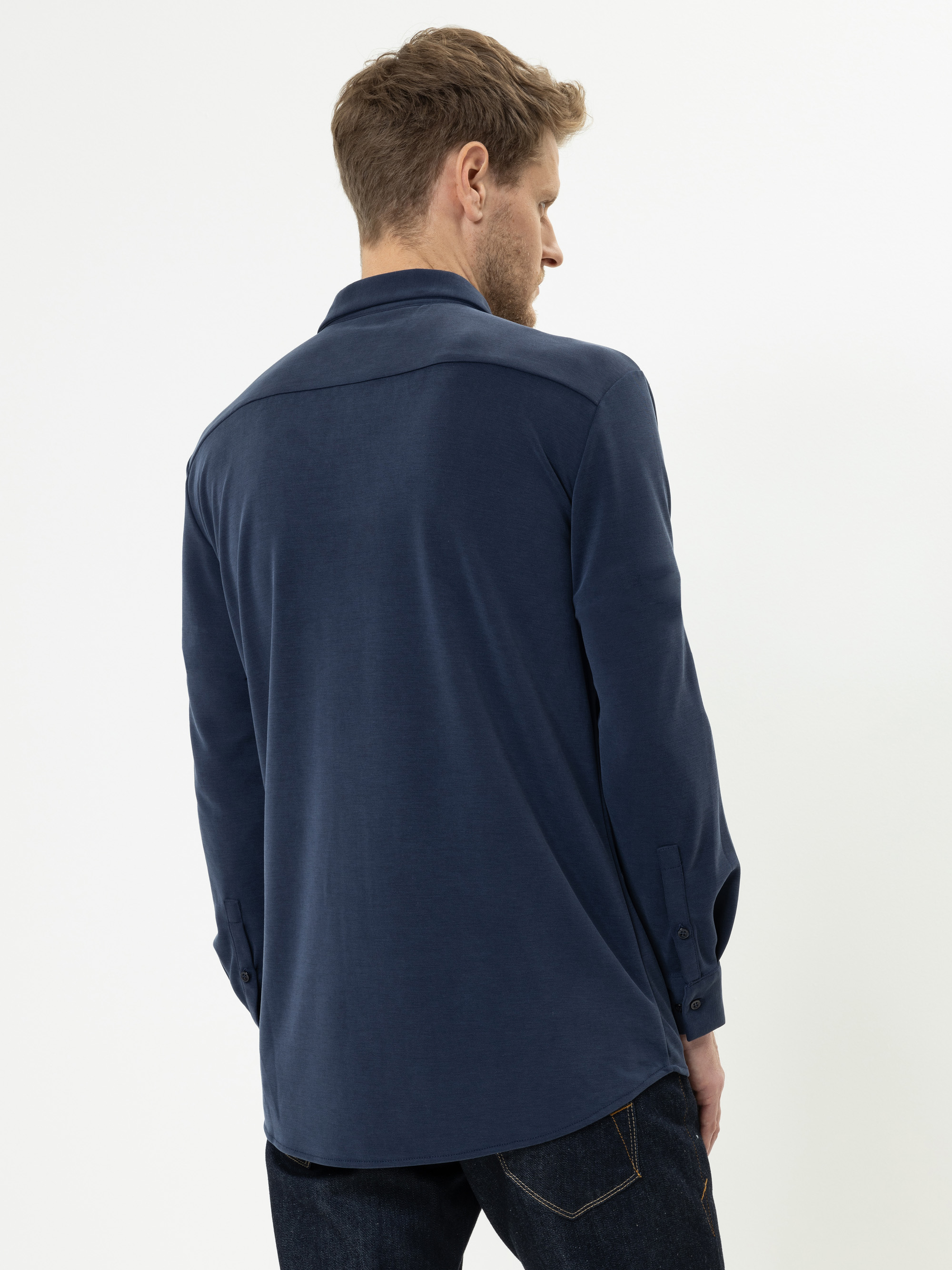 Ein Mann, der ein marineblaues, langärmeliges Hemd und dunkle Jeans trägt, steht mit dem Gesicht zur Seite und zeigt die Rückseite seines Outfits vor einem schlichten weißen Hintergrund.