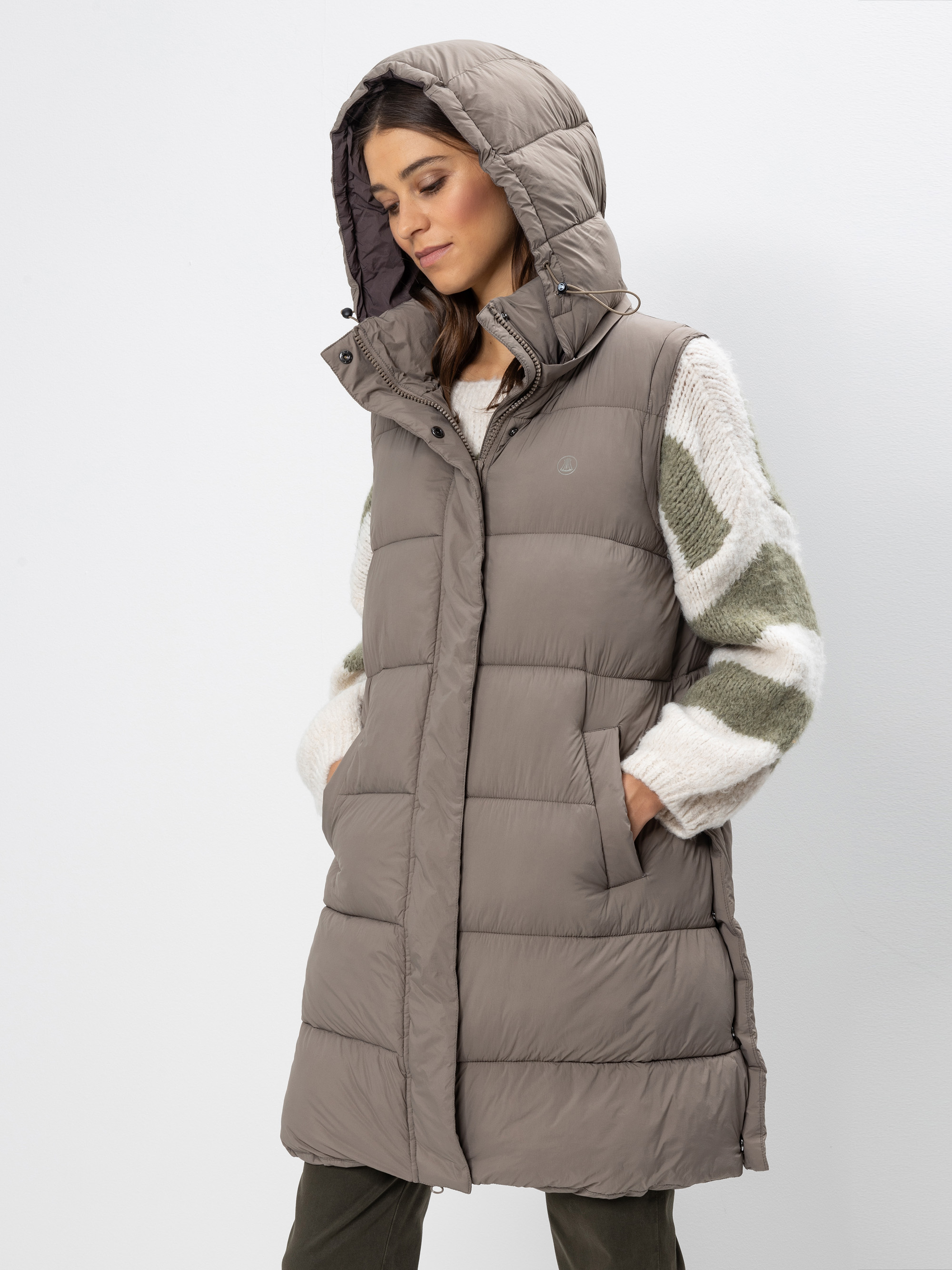 Eine Frau, die eine lange, taupefarbene Kapuzenweste über einem weiß-grünen Pullover trägt, steht mit den Händen in den Taschen da und blickt auf einen schlichten weißen Hintergrund.