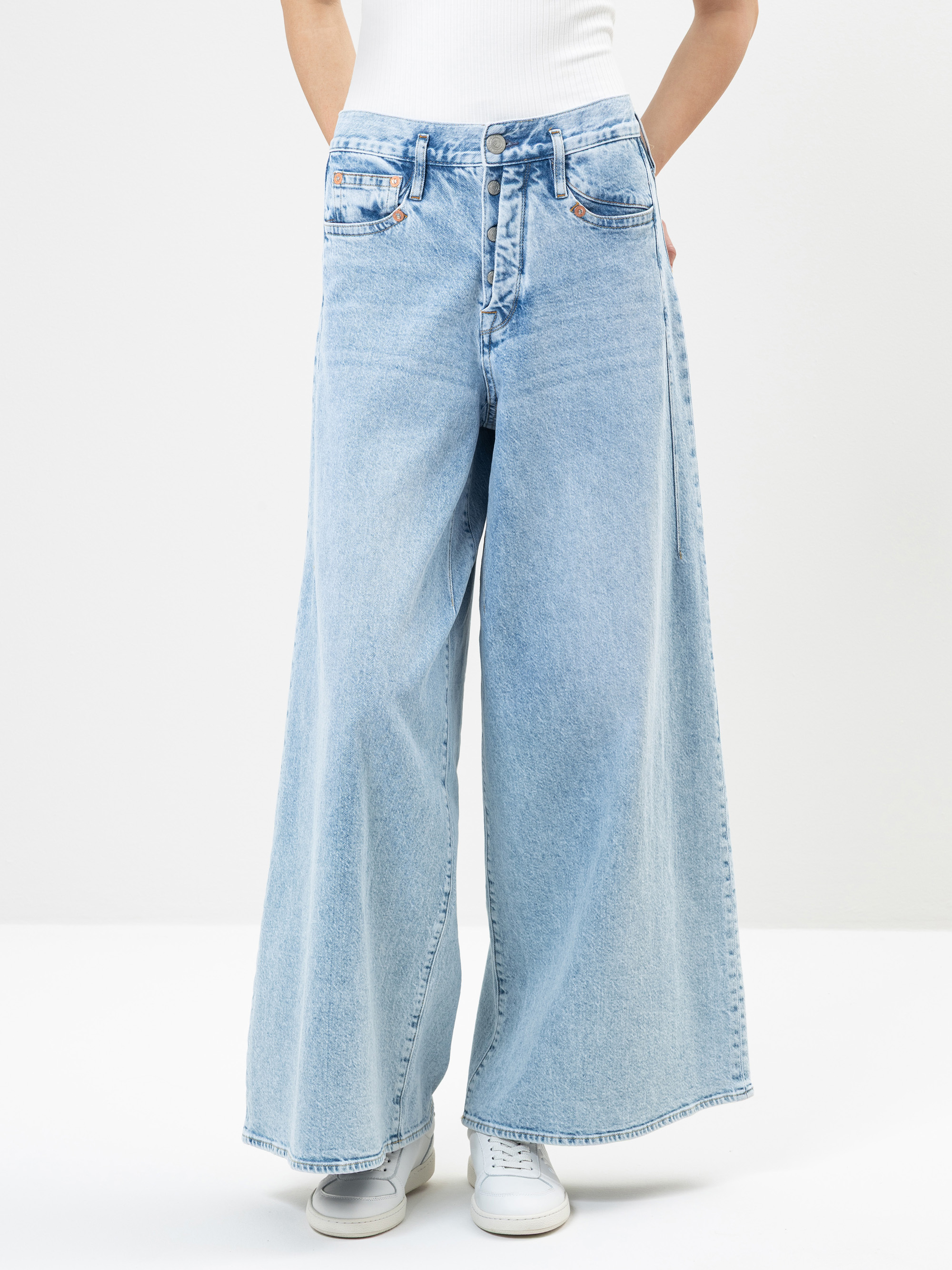 Eine Person, die hellblaue Jeans mit hoher Taille und weitem Bein, ein weißes Oberteil und weiße Turnschuhe trägt, steht mit den Händen in den Taschen vor einem schlichten weißen Hintergrund.