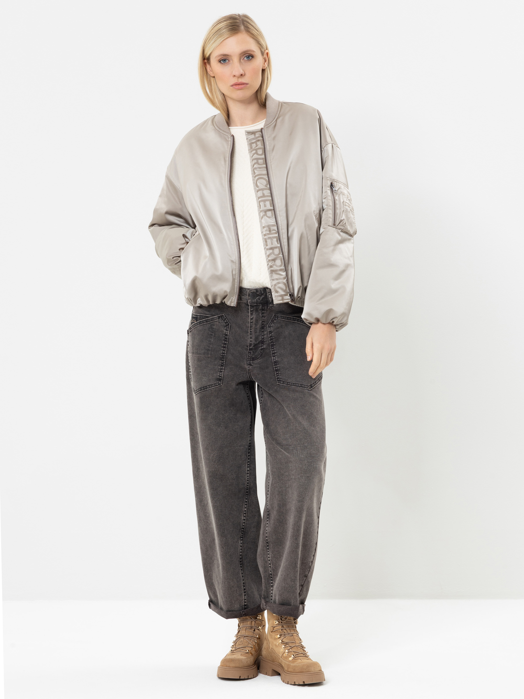 Vor einem weißen Hintergrund steht eine Frau, die eine hellbeige Bomberjacke, ein weißes Oberteil, eine weite dunkelgraue Jeans und hellbraune Schnürstiefel trägt. Sie hat glattes blondes Haar und einen ausdruckslosen Gesichtsausdruck.
