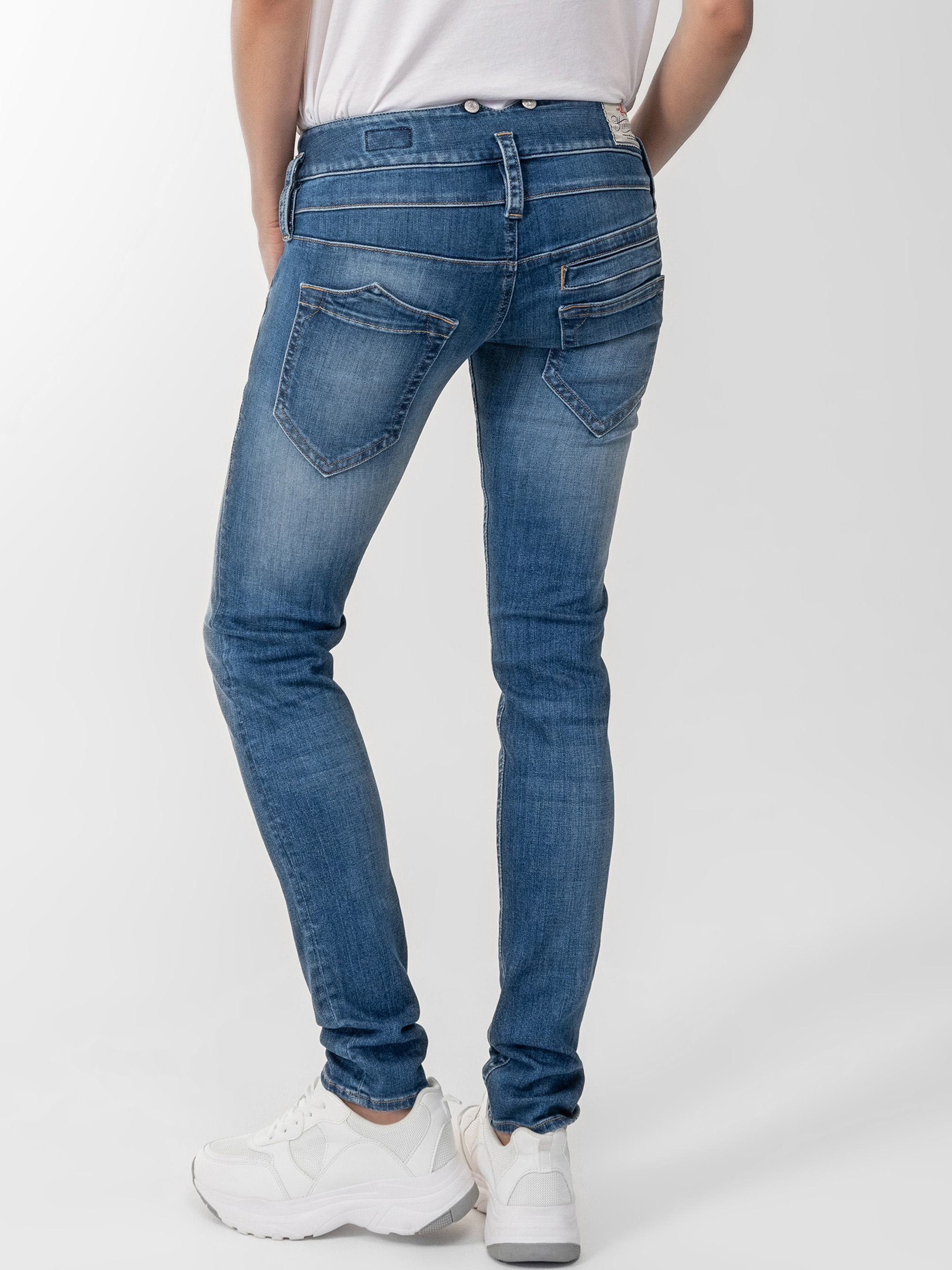 Eine Person, die eine taillierte blaue Jeans, ein weißes T-Shirt und weiße Turnschuhe trägt, steht mit dem Rücken zur Kamera vor einem schlichten, hellen Hintergrund.