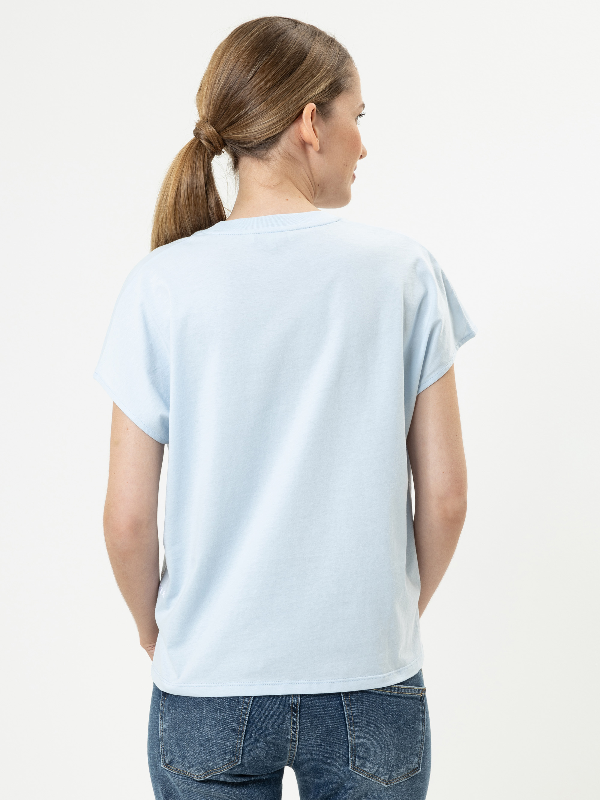 Eine Frau mit hellbraunem Haar in einem niedrigen Pferdeschwanz wird von hinten gezeigt. Sie trägt ein hellblaues, kurzärmeliges T-Shirt und blaue Jeans und steht vor einem schlichten weißen Hintergrund.