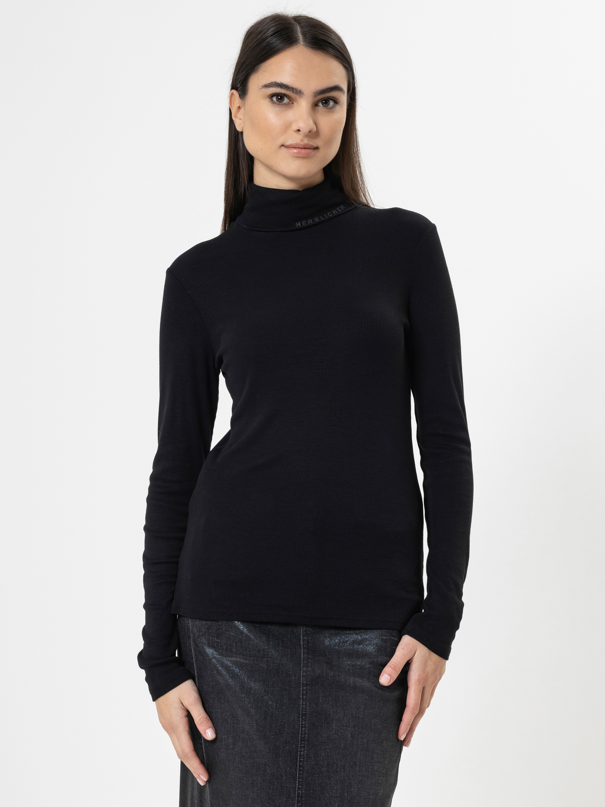 Eine Frau mit langen dunklen Haaren trägt ein schwarzes langärmeliges Rollkragenoberteil und einen dunklen Rock. Sie steht vor einem schlichten weißen Hintergrund und schaut mit neutralem Blick in die Kamera.