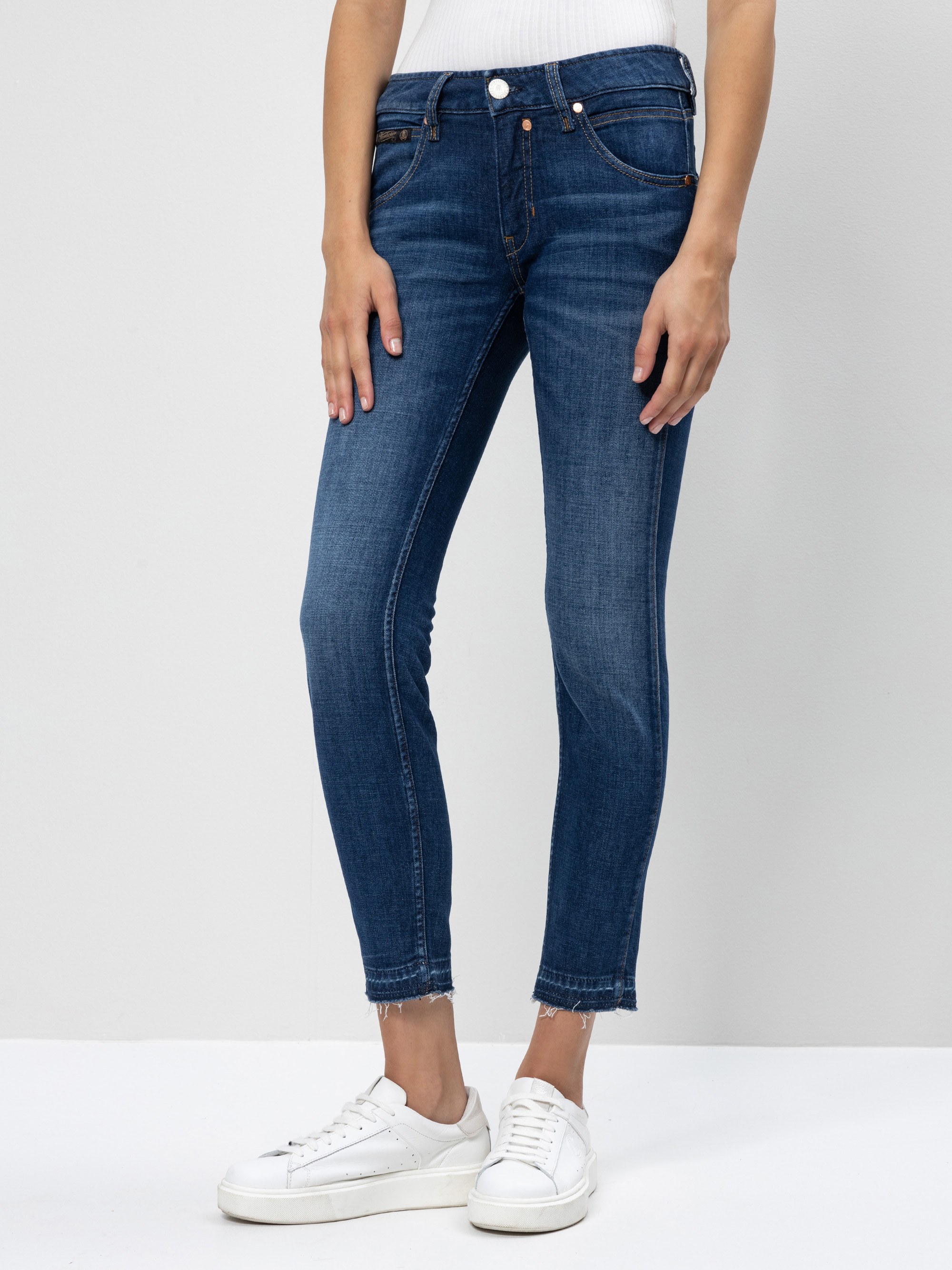 Eine Person in blauen Skinny-Jeans und weißen Turnschuhen steht vor einem einfarbigen, hellen Hintergrund und hat die Hände an die Seite gelegt. Die Jeans hat einen leicht ausgefransten Saum.