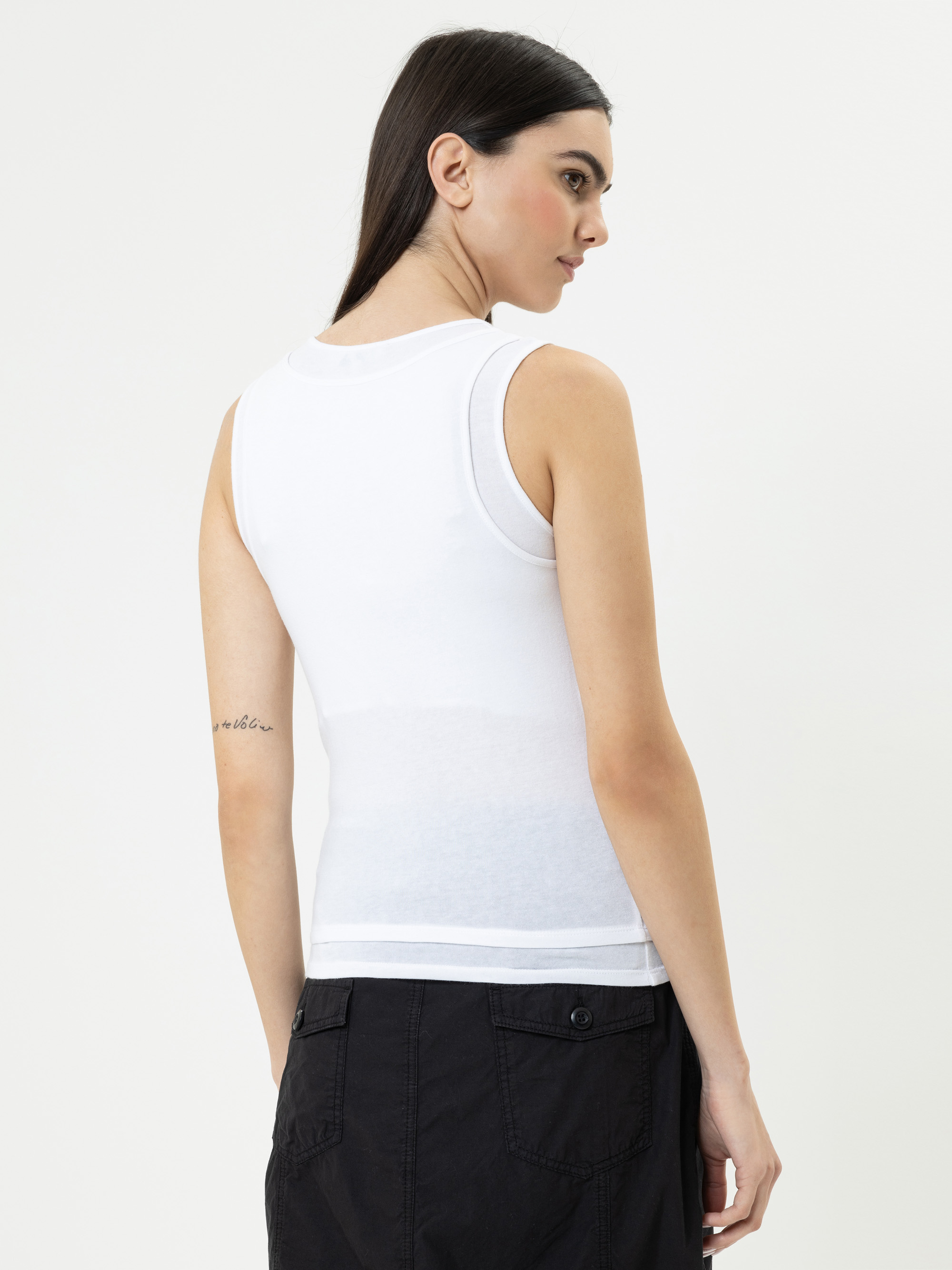 Eine Frau mit langen dunklen Haaren, die ein ärmelloses weißes Oberteil und eine schwarze Hose trägt, steht mit dem Rücken zur Kamera vor einem schlichten hellen Hintergrund. Auf ihrem linken Arm ist eine kleine Tätowierung in Schreibschrift zu sehen
