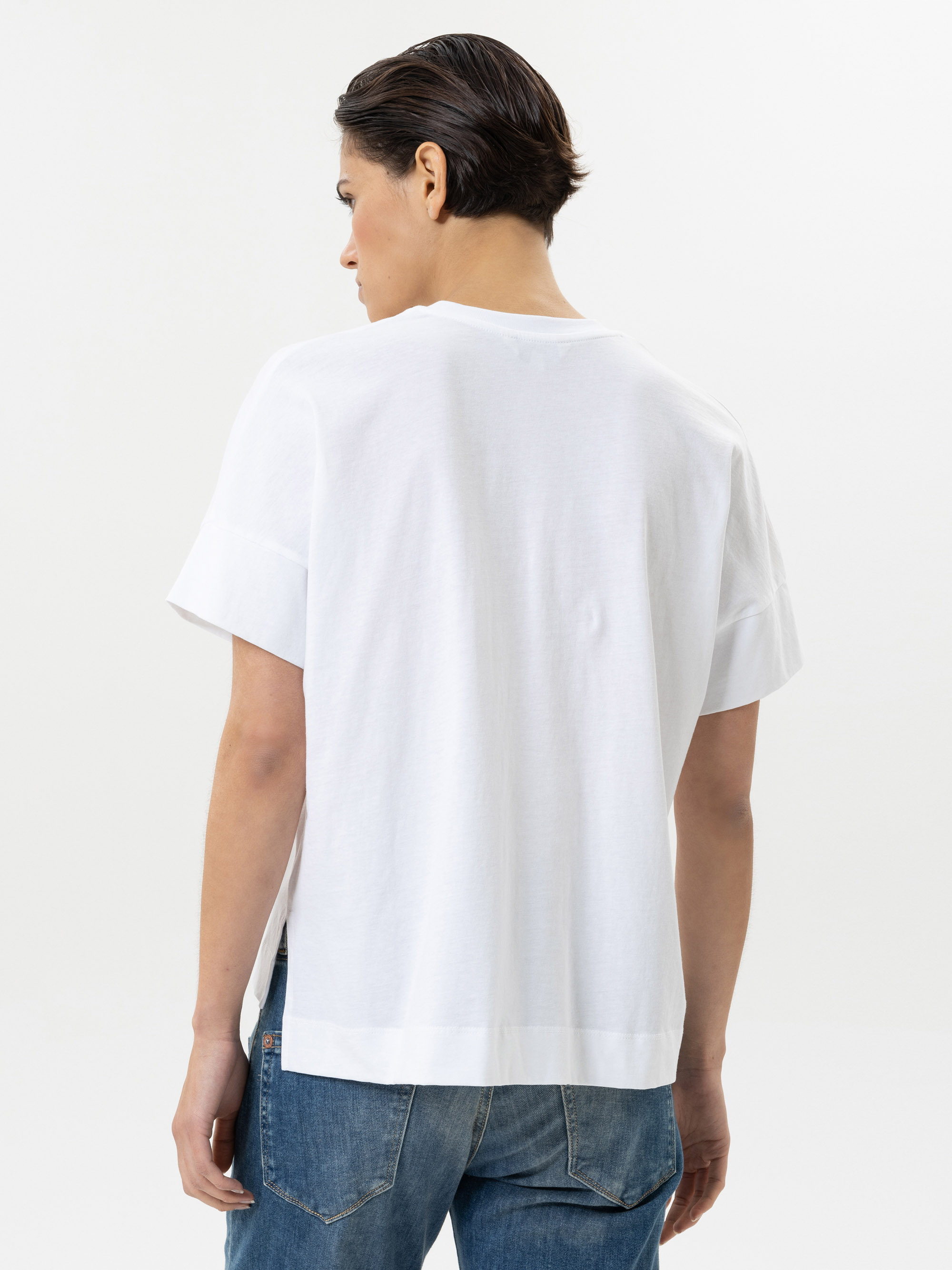 Eine Person mit kurzen dunklen Haaren, die ein lockeres weißes T-Shirt und blaue Jeans trägt, steht mit dem Rücken zur Kamera vor einem schlichten weißen Hintergrund.