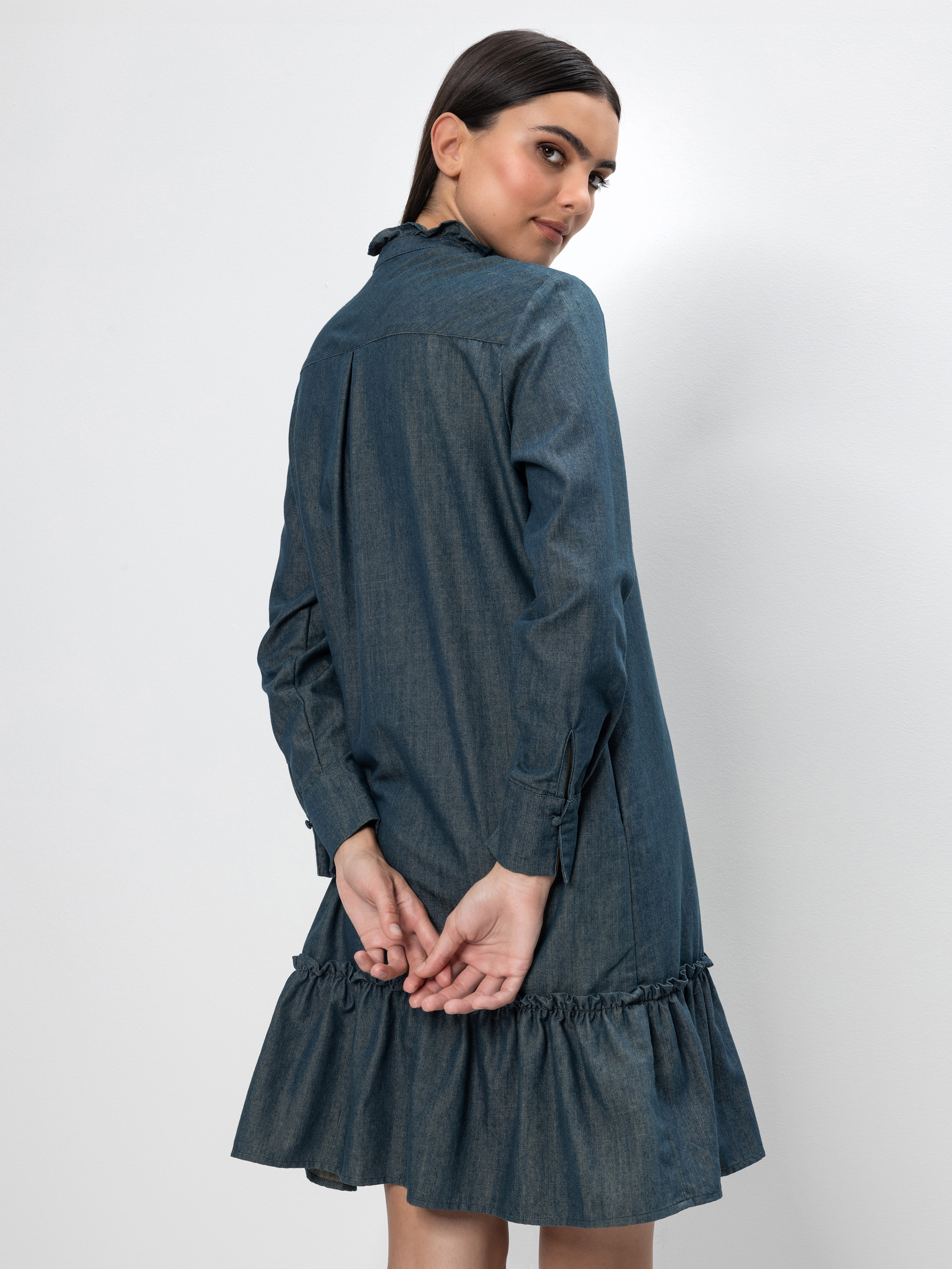 Eine Frau mit dunklem Haar und einem langärmeligen Rüschenkleid aus Jeansstoff steht mit dem Rücken zur Kamera, die Hände hinter sich verschränkt, und blickt über ihre Schulter vor einem schlichten weißen Hintergrund.