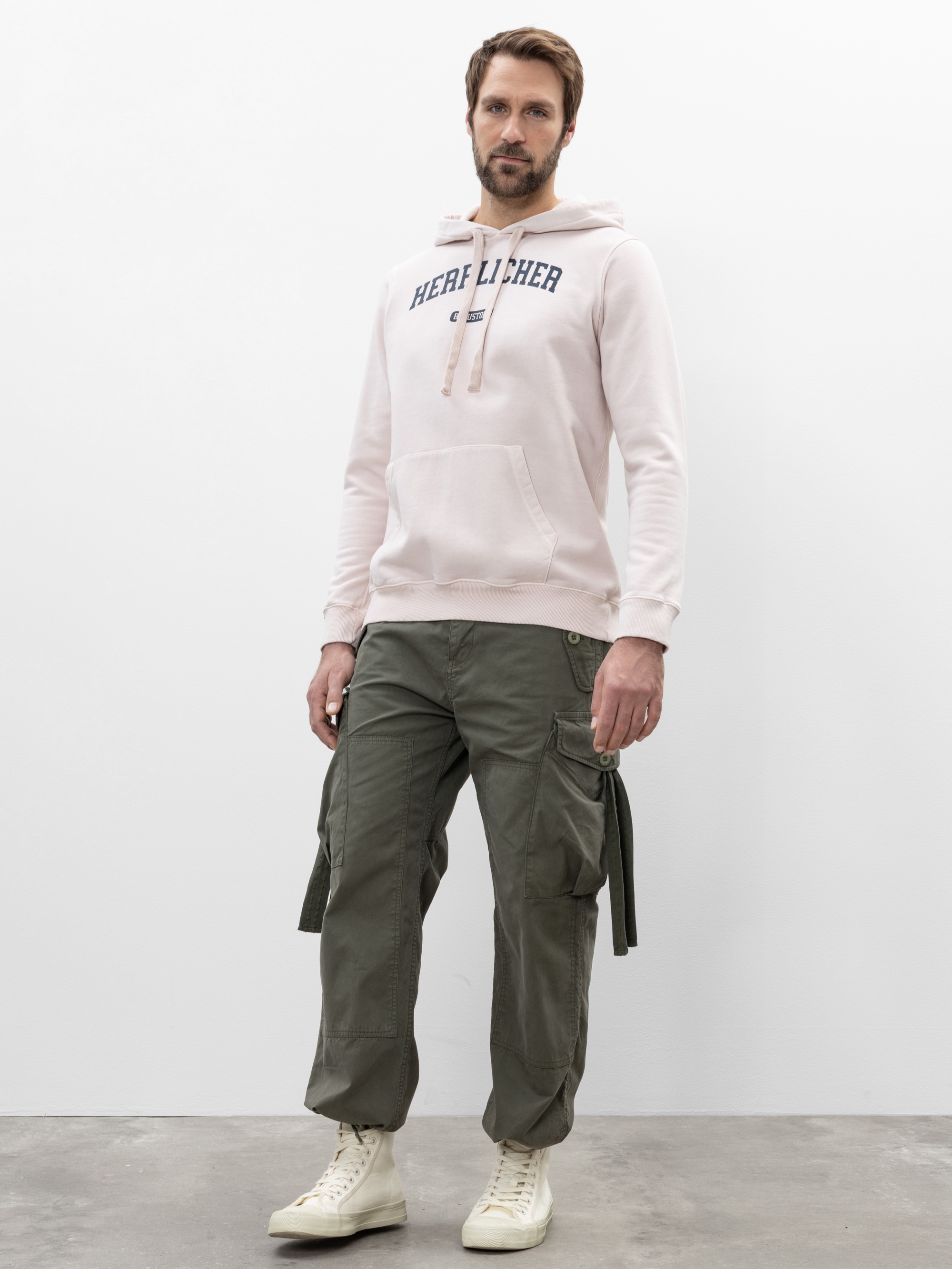 Ein Mann steht an einer schlichten weißen Wand und trägt einen hellrosa Kapuzenpulli mit dem Aufdruck HERL LICHER, eine olivgrüne Cargohose und weiße Turnschuhe mit hohen Absätzen. Er hat kurze Haare und einen Bart.