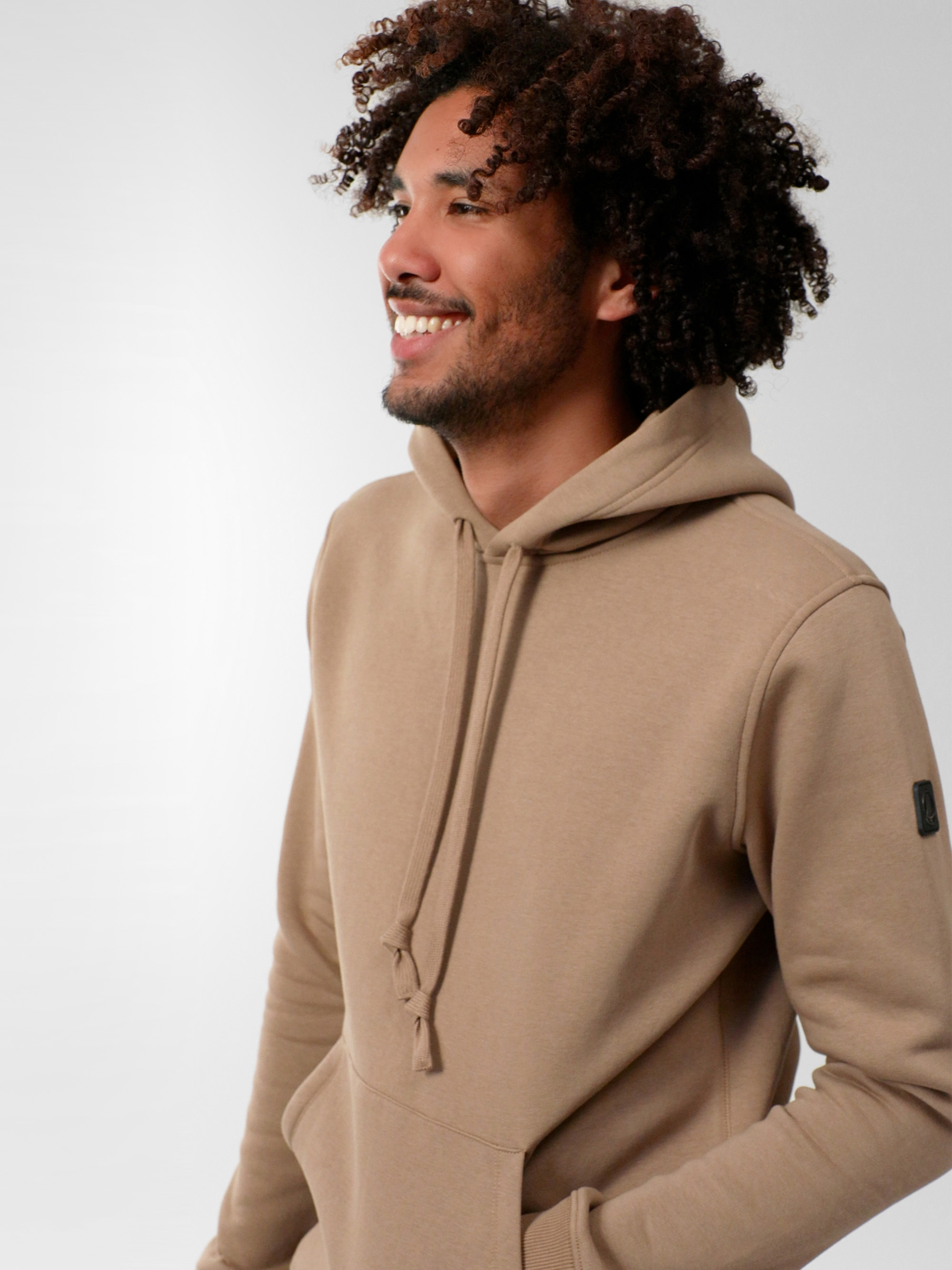 Ein lächelnder Mann mit lockigem Haar und hellbraunem Kapuzenpulli steht vor einem schlichten, hellen Hintergrund und blickt in die Ferne.