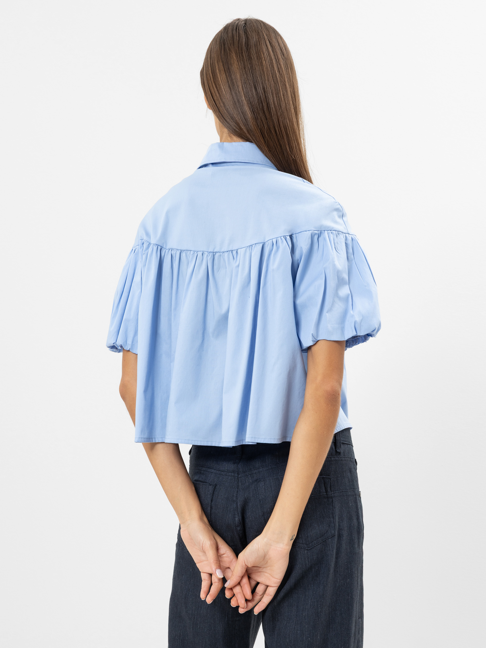 Eine Frau mit langen braunen Haaren steht mit dem Gesicht nach hinten und trägt eine hellblaue, kurzärmelige, locker sitzende Bluse und eine dunkle Hose. Sie hält ihre Hände hinter ihrem Rücken vor einem schlichten weißen Hintergrund.