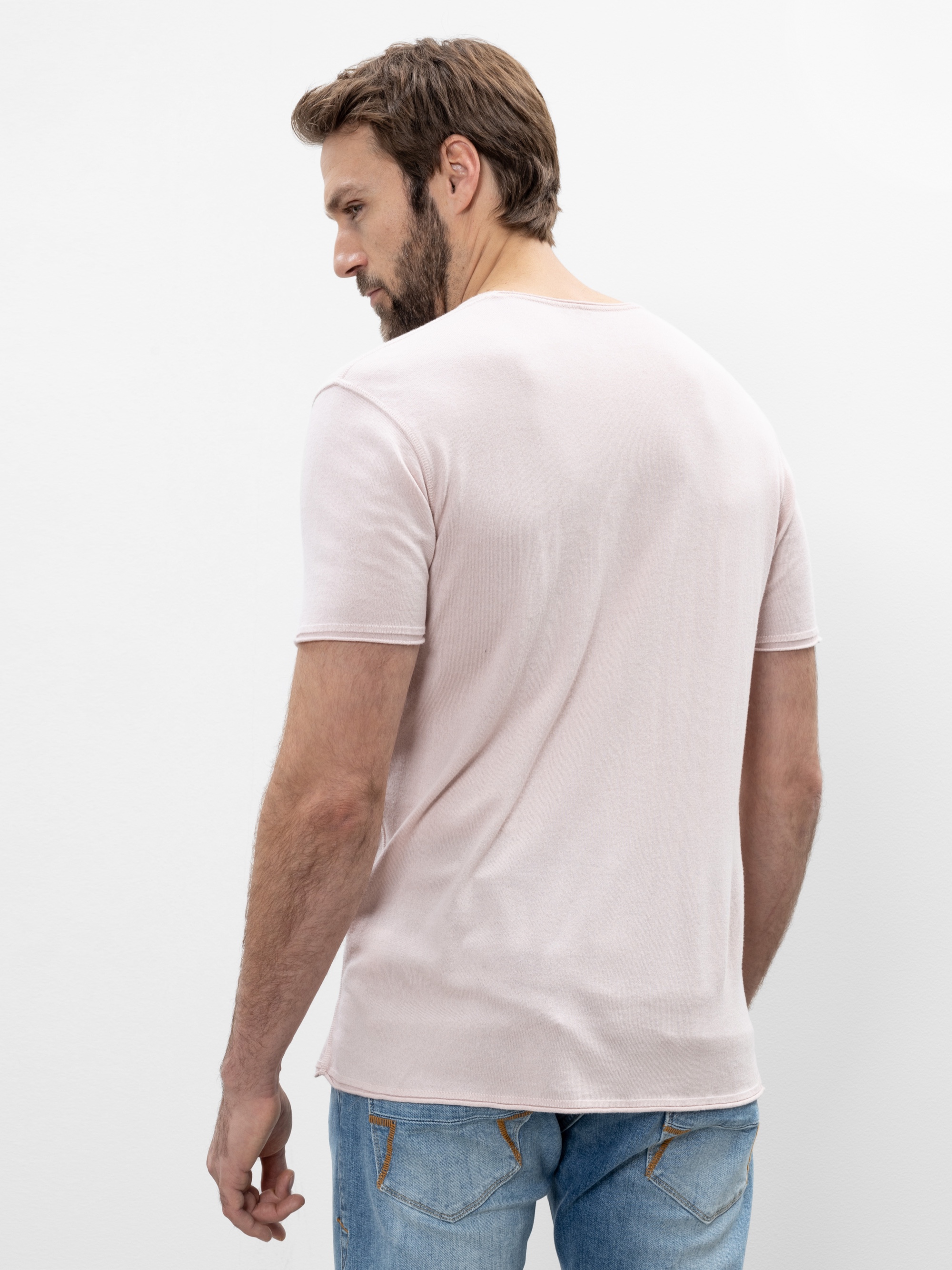 Ein Mann mit kurzen braunen Haaren und Bart steht mit dem Rücken zur Kamera, trägt ein hellrosa T-Shirt und blaue Jeans vor einem schlichten weißen Hintergrund.