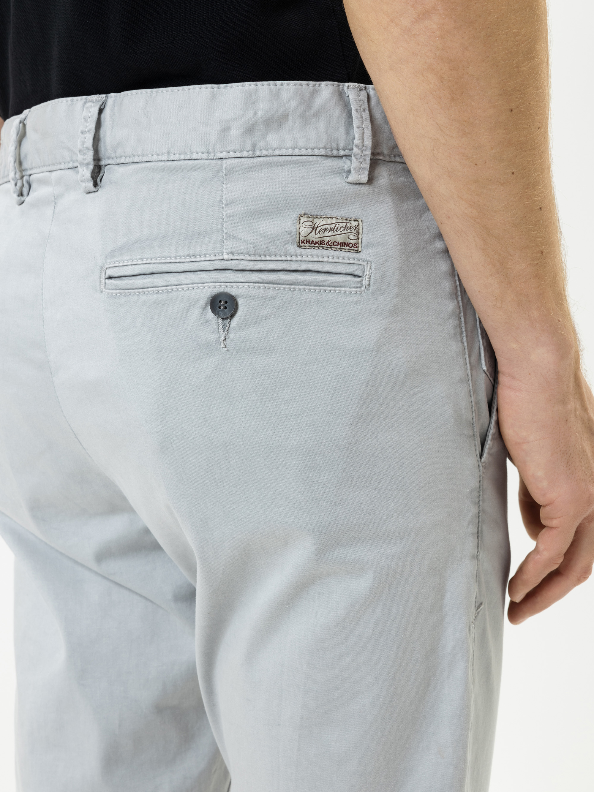 Eine Person trägt eine hellgraue Hose mit einer Gesäßtasche, die mit einem Knopf und einem kleinen rechteckigen Stoffetikett über der Tasche versehen ist. Der rechte Arm der Person ist sichtbar, und sie trägt ein schwarzes Hemd.