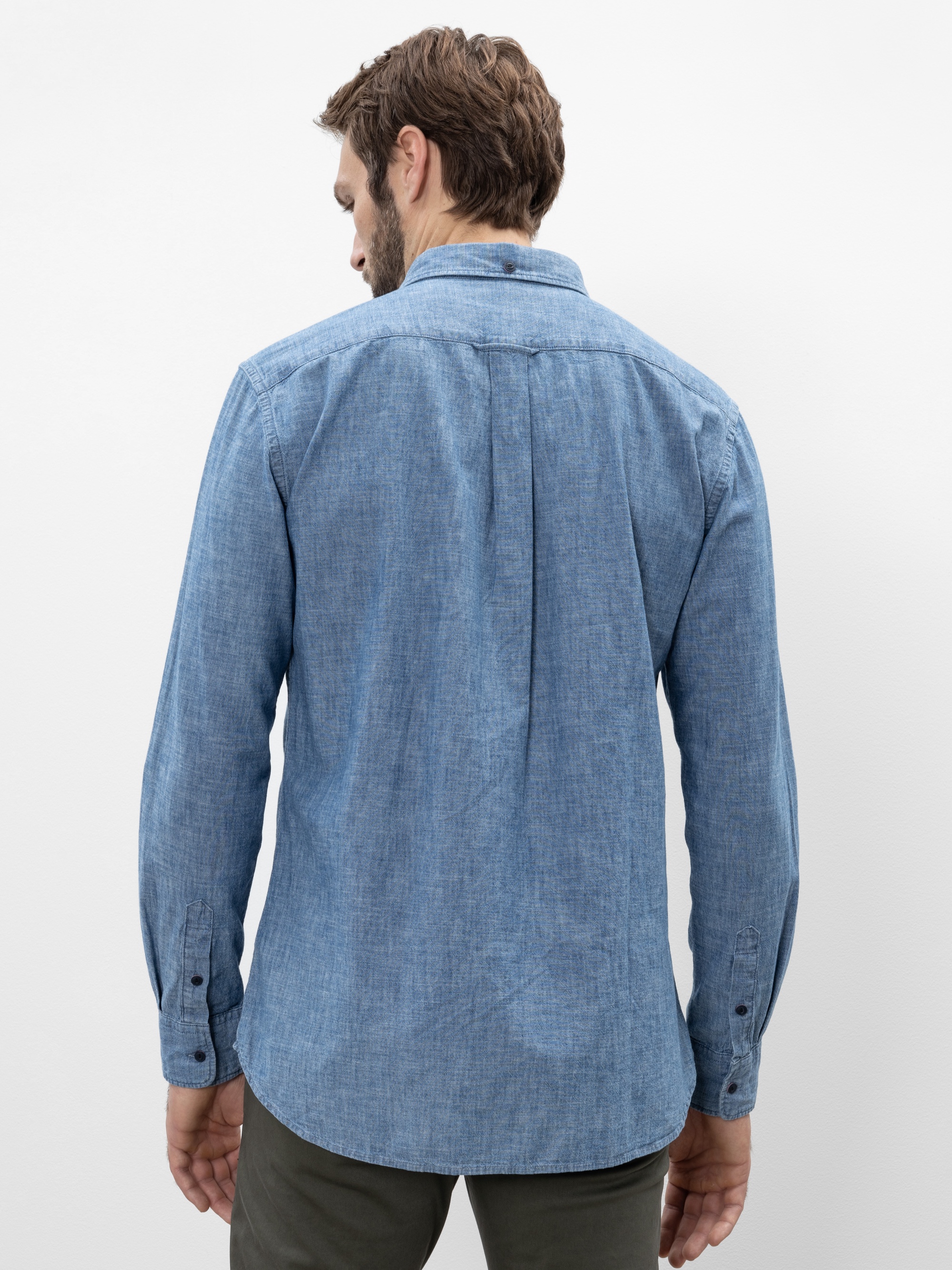 Ein Mann mit kurzen braunen Haaren steht mit dem Rücken zur Kamera. Er trägt ein hellblaues, langärmeliges Button-up-Hemd und eine dunkle Hose vor einem schlichten weißen Hintergrund.