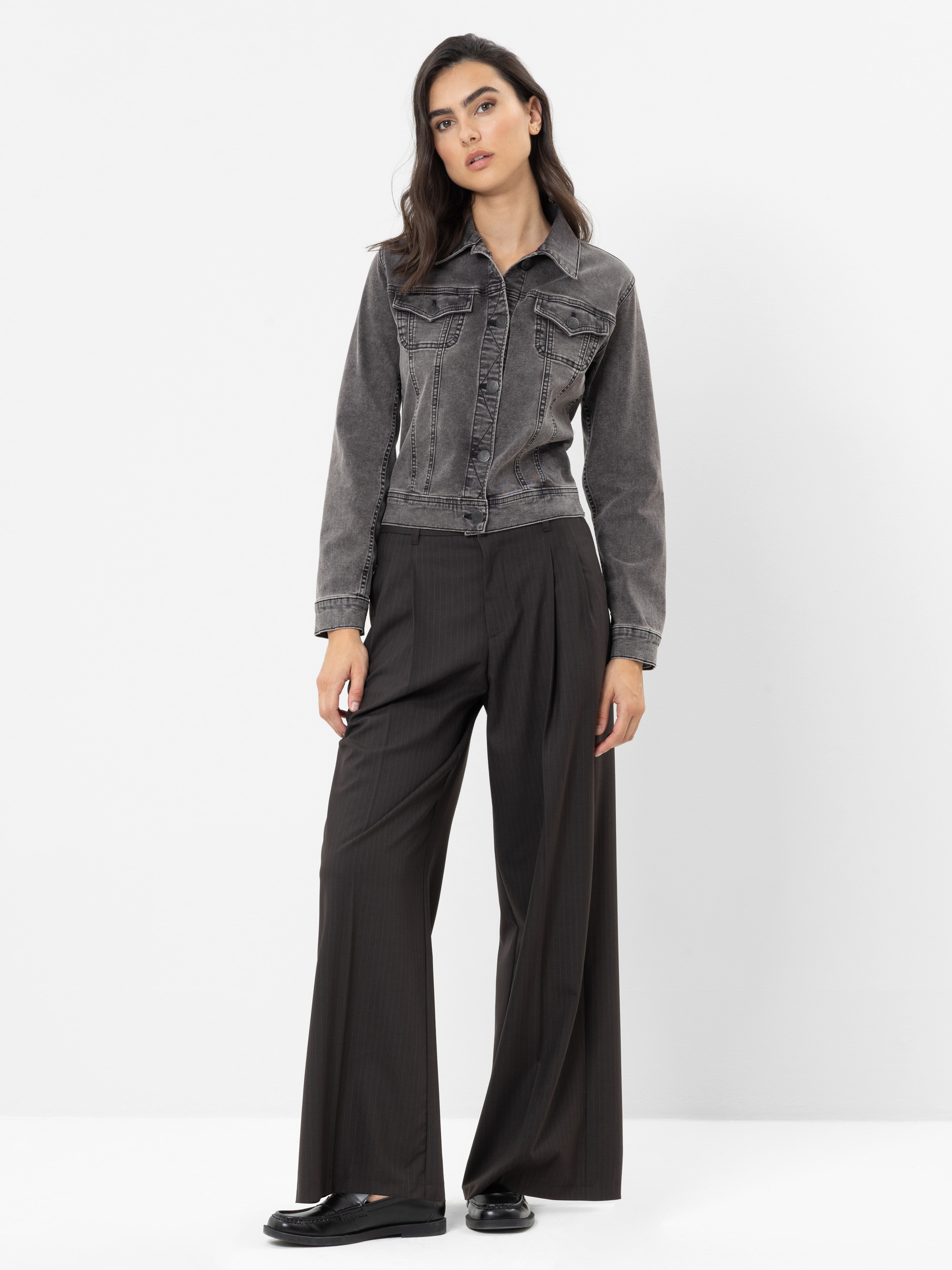 Vor einem weißen Hintergrund steht eine Frau, die eine graue Jeansjacke, eine weite schwarze Nadelstreifenhose und schwarze Schuhe trägt.