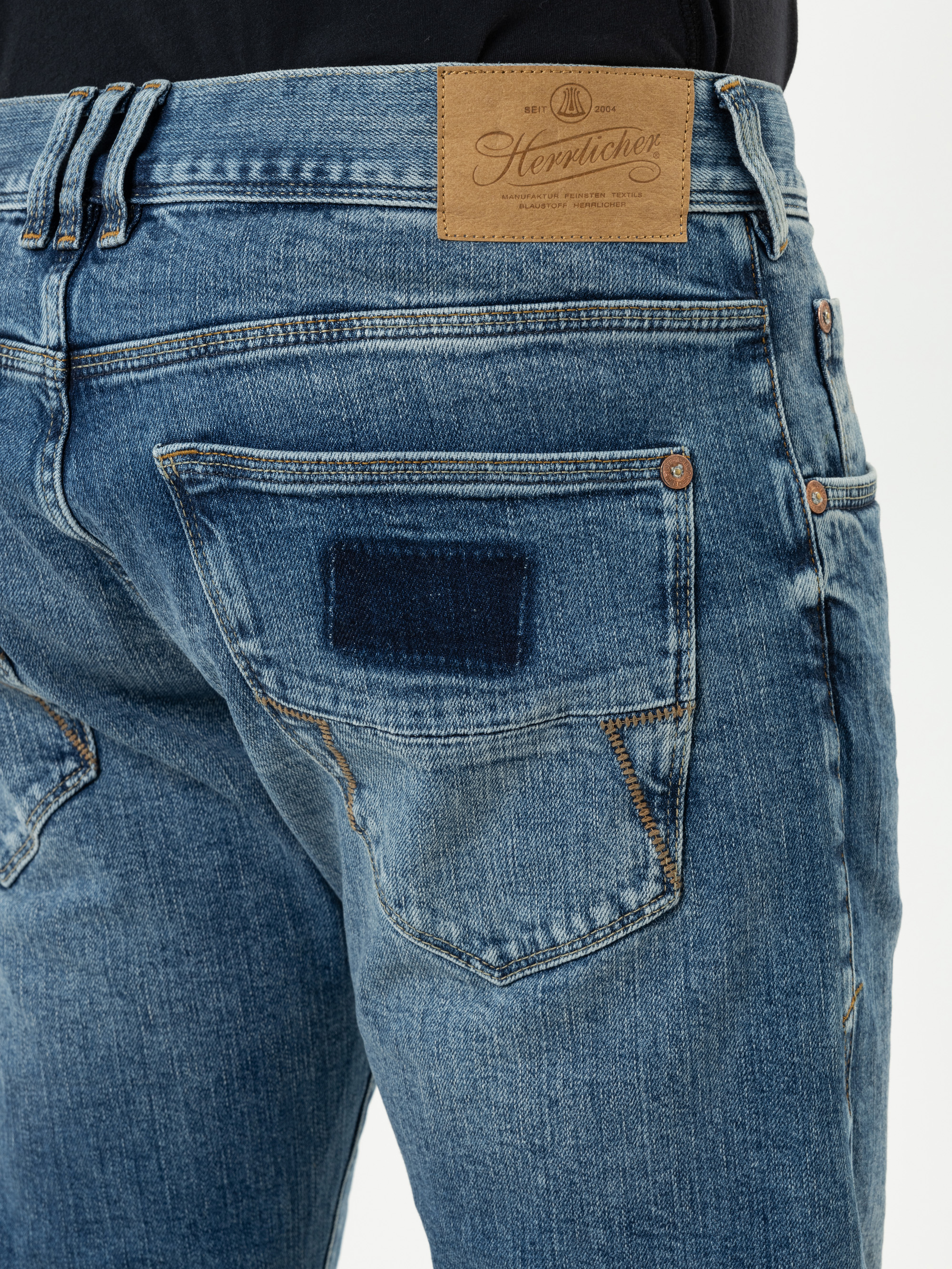 Nahaufnahme des Rückens einer Person, die eine blaue Denim-Jeans mit einem braunen Lederaufnäher der Marke Herrlicher und einem dunkelblauen rechteckigen Aufnäher auf der Gesäßtasche trägt.