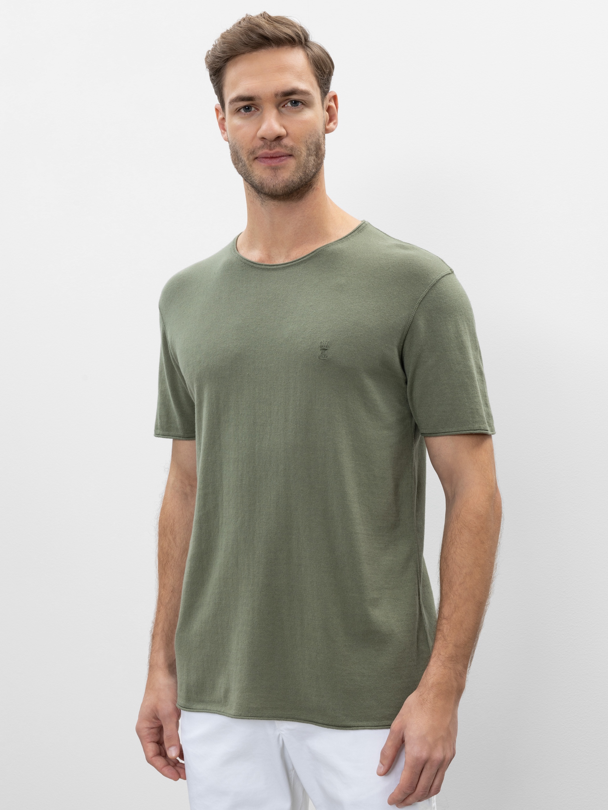 Ein Mann mit kurzen braunen Haaren und gestutztem Bart trägt ein schlichtes olivgrünes T-Shirt und eine weiße Hose. Er steht vor einem schlichten hellen Hintergrund und blickt mit einem leichten Lächeln in die Kamera.