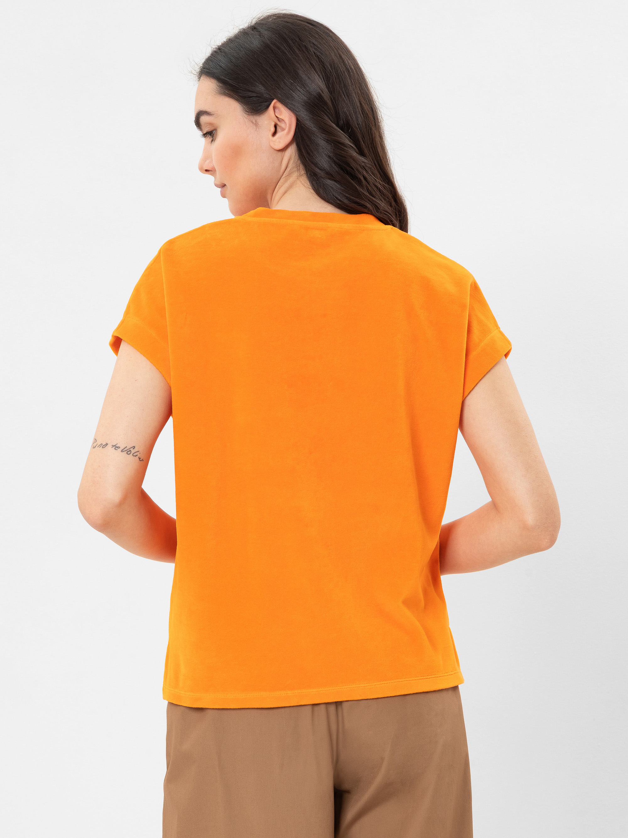 Eine Frau mit langen dunklen Haaren wird von hinten gezeigt. Sie trägt ein orangefarbenes kurzärmeliges Oberteil und eine braune Hose. Sie hat eine Tätowierung auf ihrem rechten Unterarm. Der Hintergrund ist einfarbig weiß.