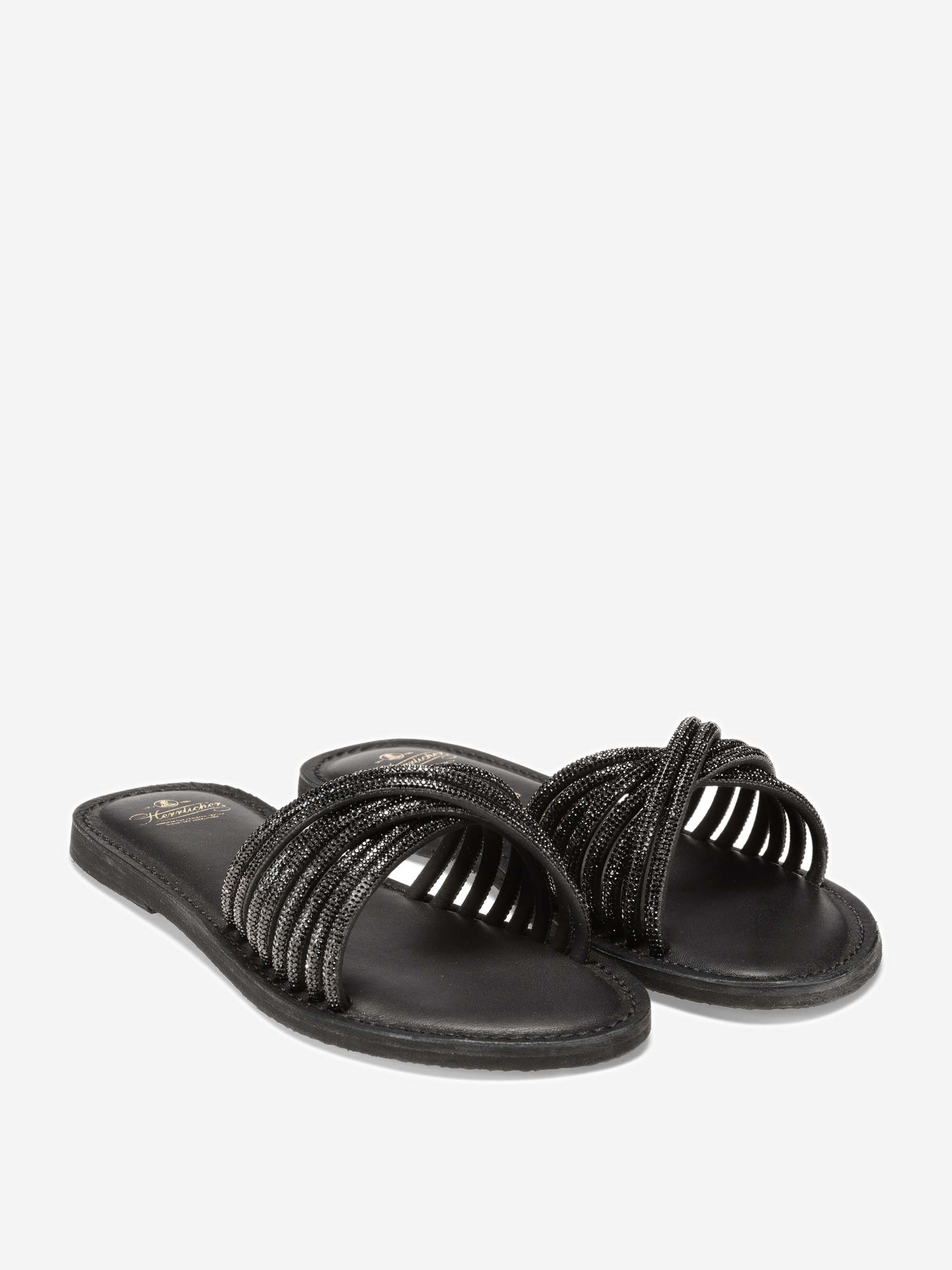 Ein Paar schwarzer, flacher Slide-Sandalen mit sich kreuzenden, perlenbesetzten Riemen und offener Zehenpartie auf weißem Hintergrund.
