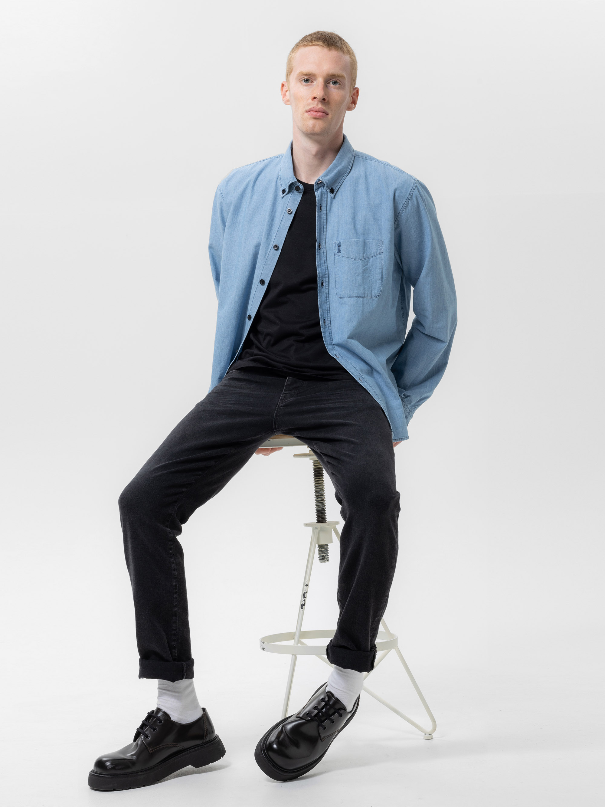 Eine Person mit kurzen hellen Haaren sitzt auf einem weißen Hocker und trägt ein hellblaues Button-up-Hemd über einem schwarzen T-Shirt, schwarze Jeans, weiße Socken und schwarze Schuhe vor einem schlichten weißen Hintergrund.