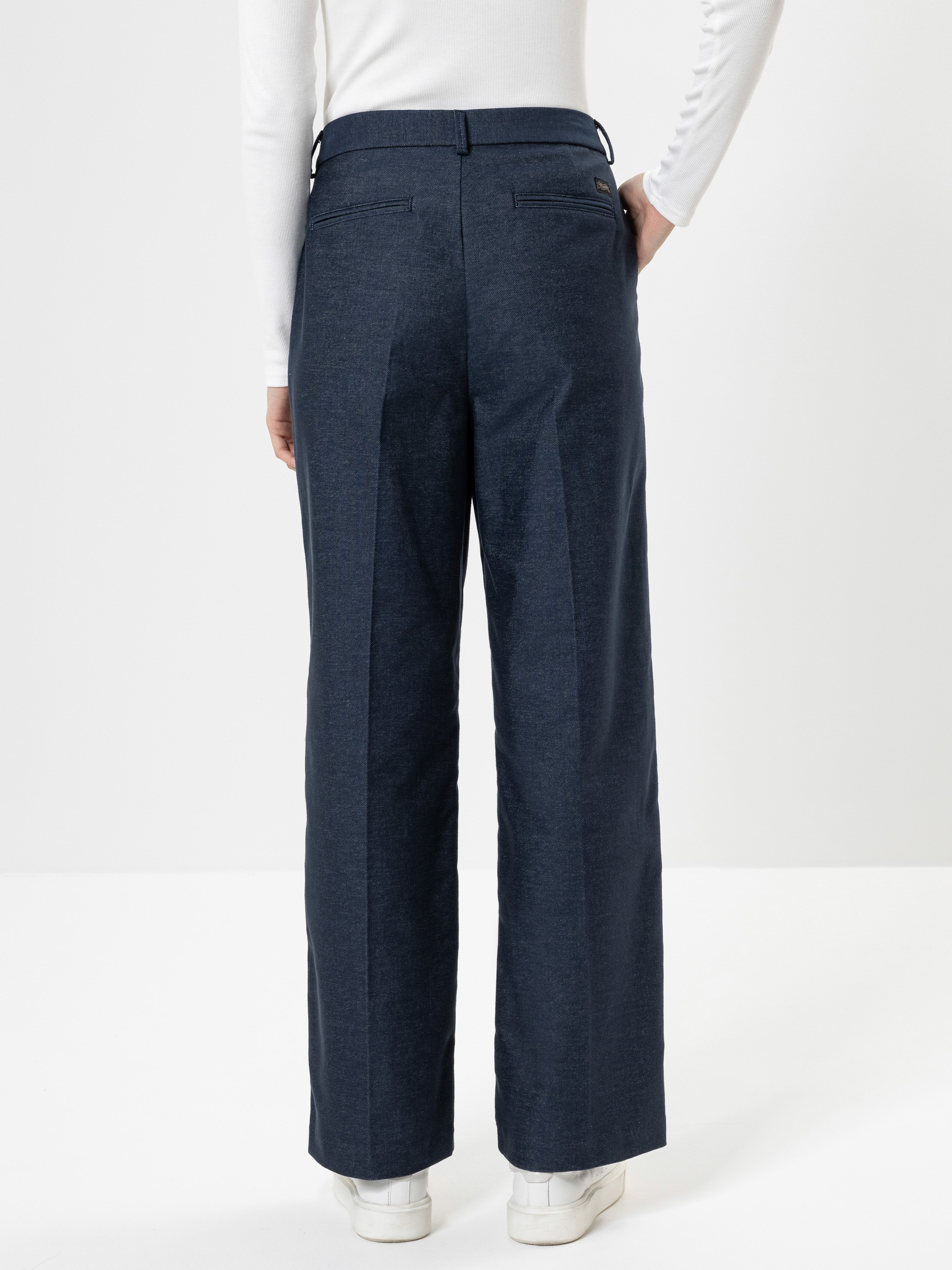 Eine Person, die eine dunkelblaue Hose mit weitem Bein, ein weißes langärmeliges Oberteil und weiße Turnschuhe trägt, wird von hinten vor einem einfarbig weißen Hintergrund gezeigt.