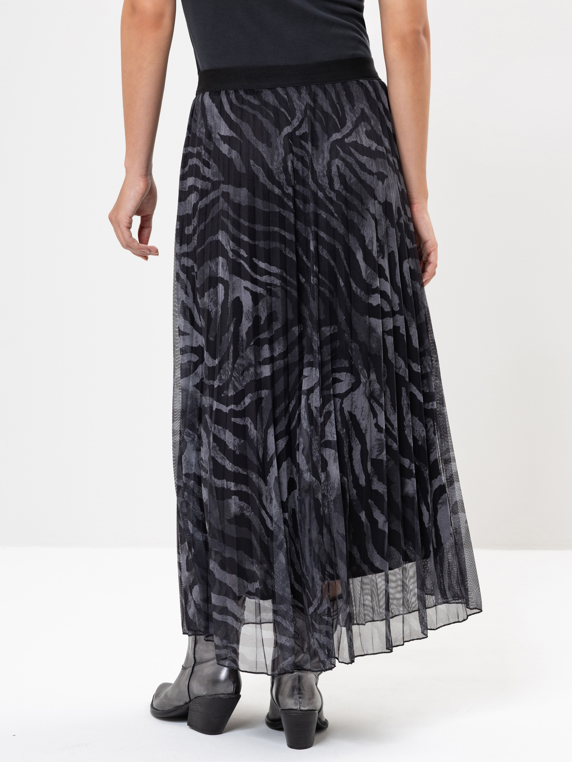 Eine Person, die einen schwarz-grauen Faltenrock mit Zebramuster und grauen Ankle Boots trägt. Das Bild ist von hinten aufgenommen und zeigt die fließende Textur des Rocks.