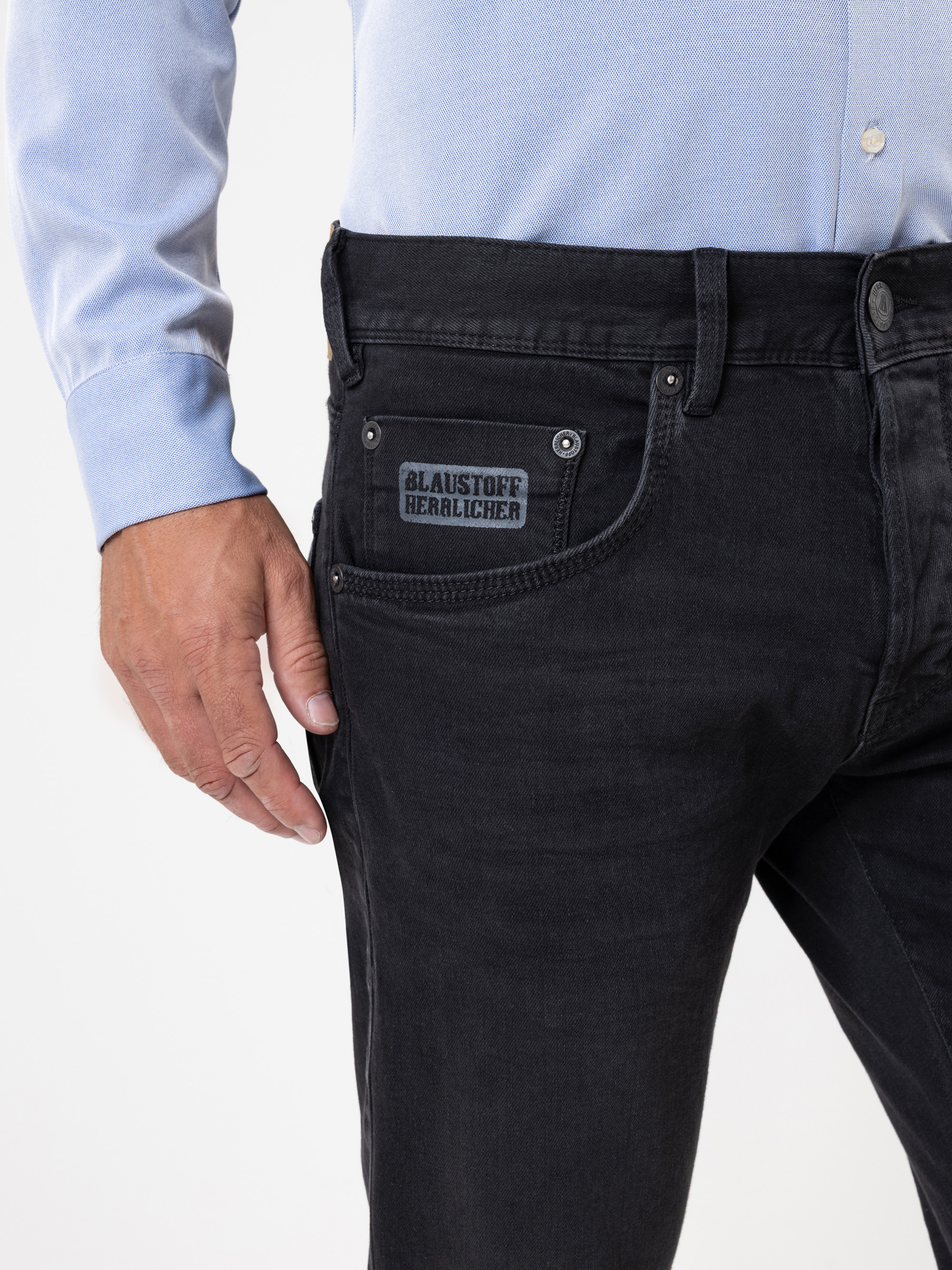 Eine Person, die ein hellblaues Button-up-Hemd und eine schwarze Jeans mit einem Etikett an der Tasche trägt, auf dem "BLAUSTOFF HERZLICHER" steht. Nur der untere Teil des Oberkörpers und der linke Arm sind sichtbar.