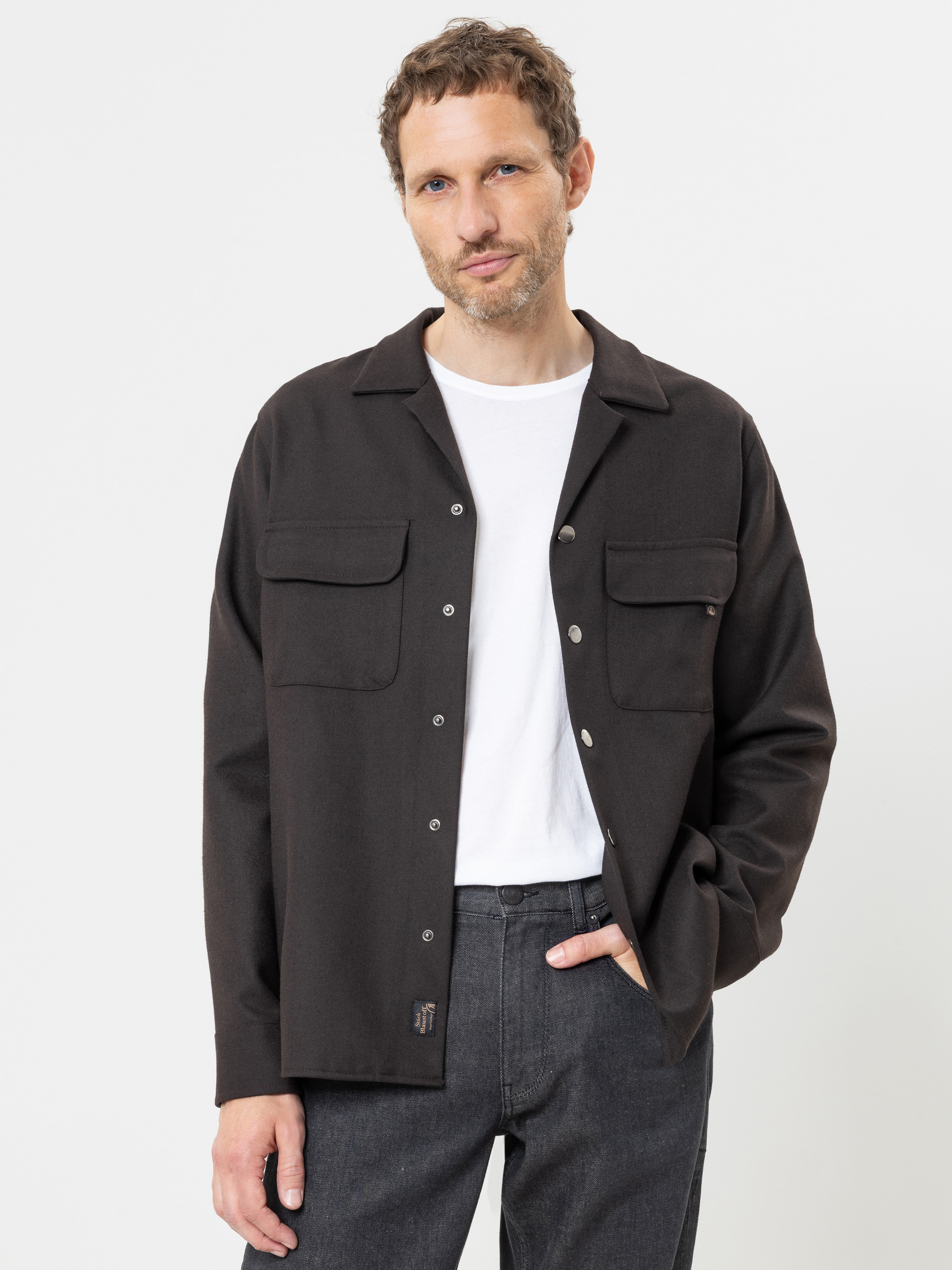 Ein Mann mit kurzen Locken trägt ein schwarzes Hemd mit Knöpfen über einem weißen T-Shirt und eine dunkle Jeans; er steht vor einem schlichten weißen Hintergrund und hat eine Hand in der Hosentasche.
