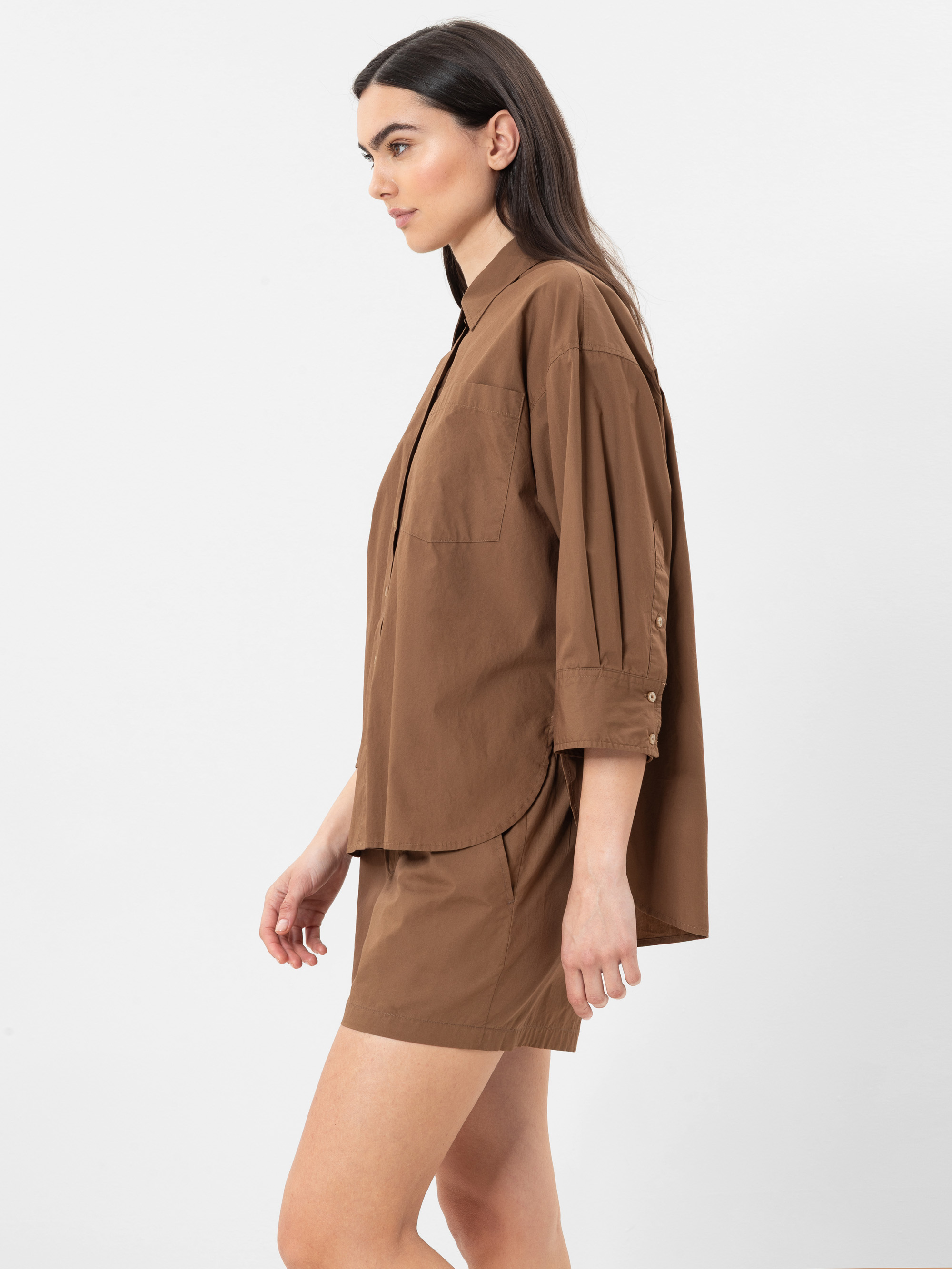 Eine Frau in einem braunen Button-up-Hemd mit hochgekrempelten Ärmeln und passenden braunen Shorts steht im Profil vor einem schlichten weißen Hintergrund.