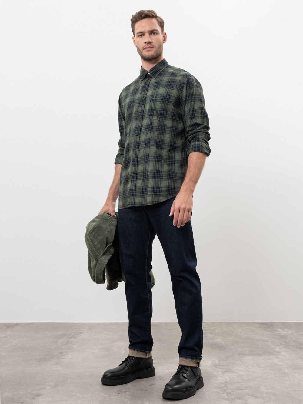 Ein Mann steht in einem Innenraum vor einer schlichten weißen Wand. Er trägt ein grün kariertes Button-up-Hemd, dunkle Jeans und schwarze Schuhe. Er hält eine grüne Jacke in seiner linken Hand und blickt selbstbewusst in die Kamera.