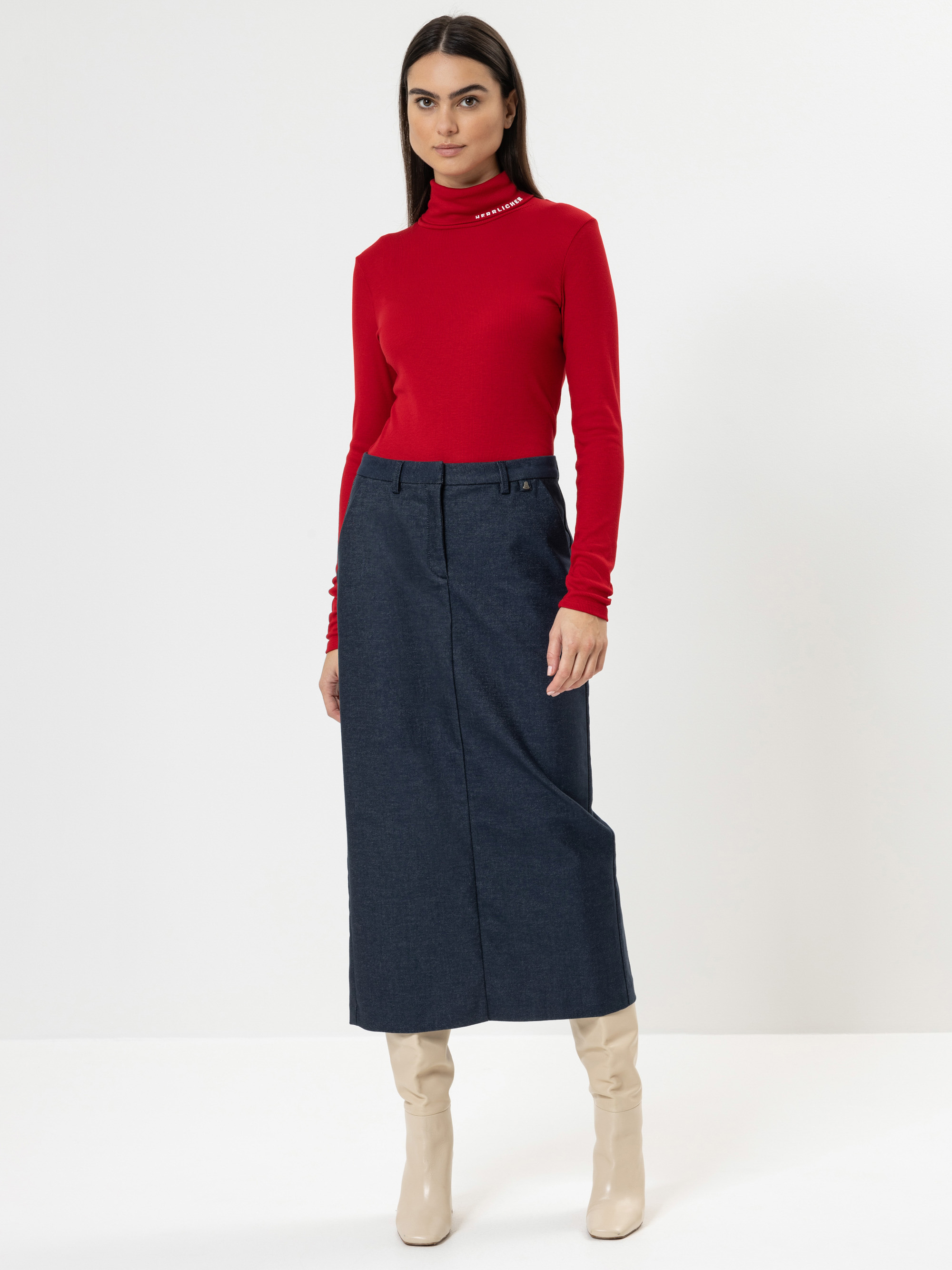 Eine Frau steht vor einem weißen Hintergrund und trägt ein rotes langärmeliges Rollkragenoberteil, einen hochtaillierten Midirock aus dunklem Jeansstoff und kniehohe cremefarbene Stiefel. Sie hat glattes dunkles Haar und einen neutralen Gesichtsausdruc