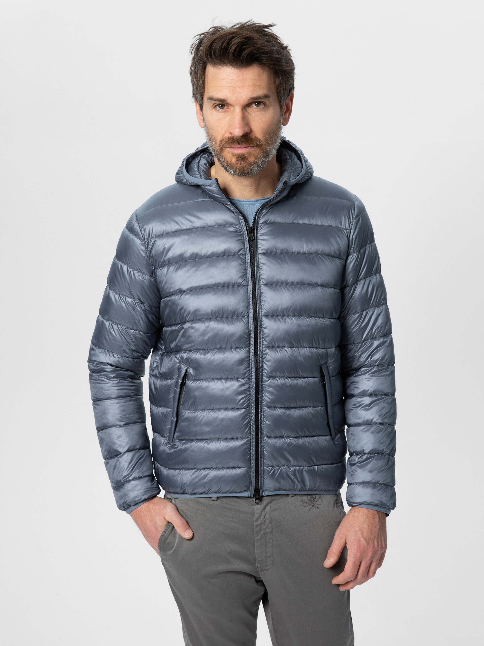 Ein Mann mit braunem Haar und Bart trägt eine glänzende, graue Pufferjacke mit Kapuze, ein hellgraues Hemd und eine graue Hose und steht vor einem schlichten weißen Hintergrund.
