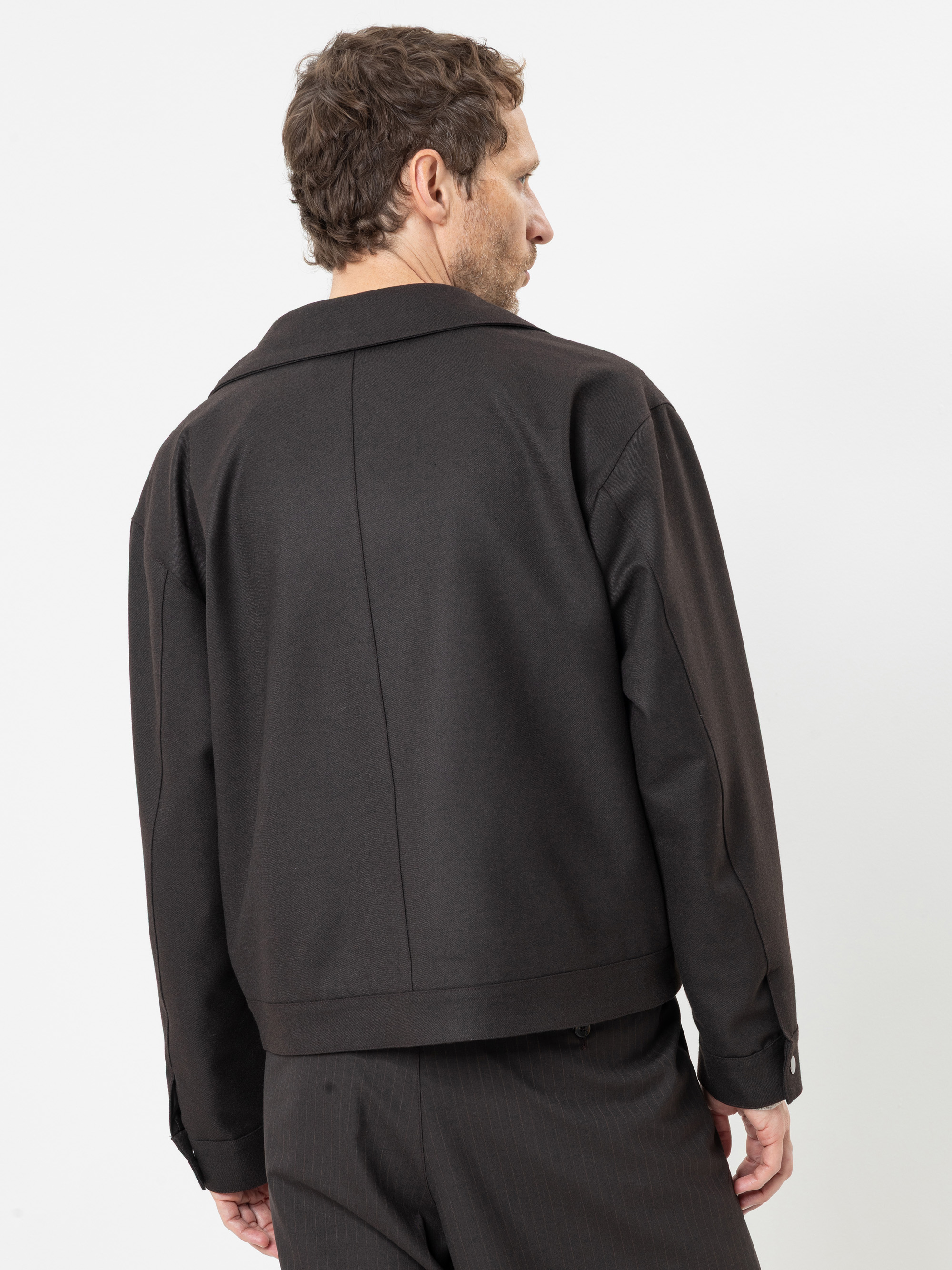 Ein Mann mit kurzen braunen Haaren ist von hinten zu sehen; er trägt eine schwarze Jacke und eine dunkle Hose und steht vor einem schlichten weißen Hintergrund.