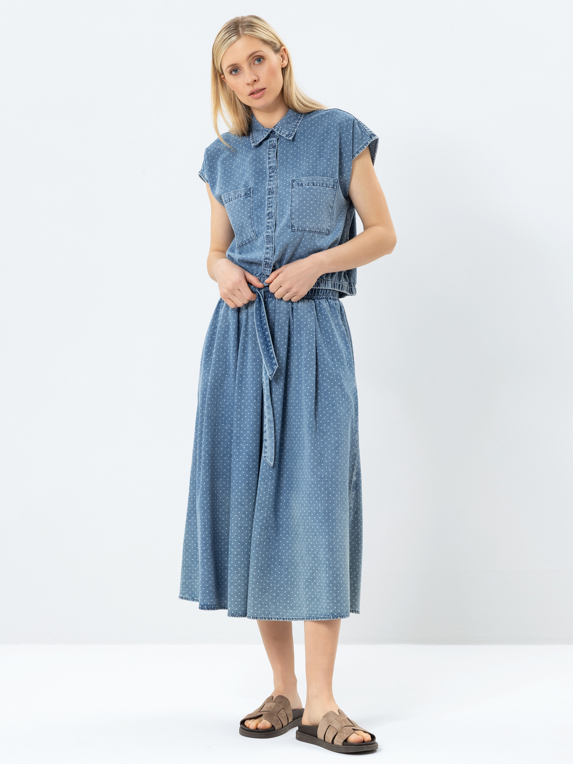 Eine Frau steht vor einem unifarbenen Hintergrund und trägt ein ärmelloses, hellblaues Jeanshemd mit Vordertaschen und einen dazu passenden Midirock mit hoher Taille, der in der Taille gebunden wird. Außerdem trägt sie braune Sandalen.