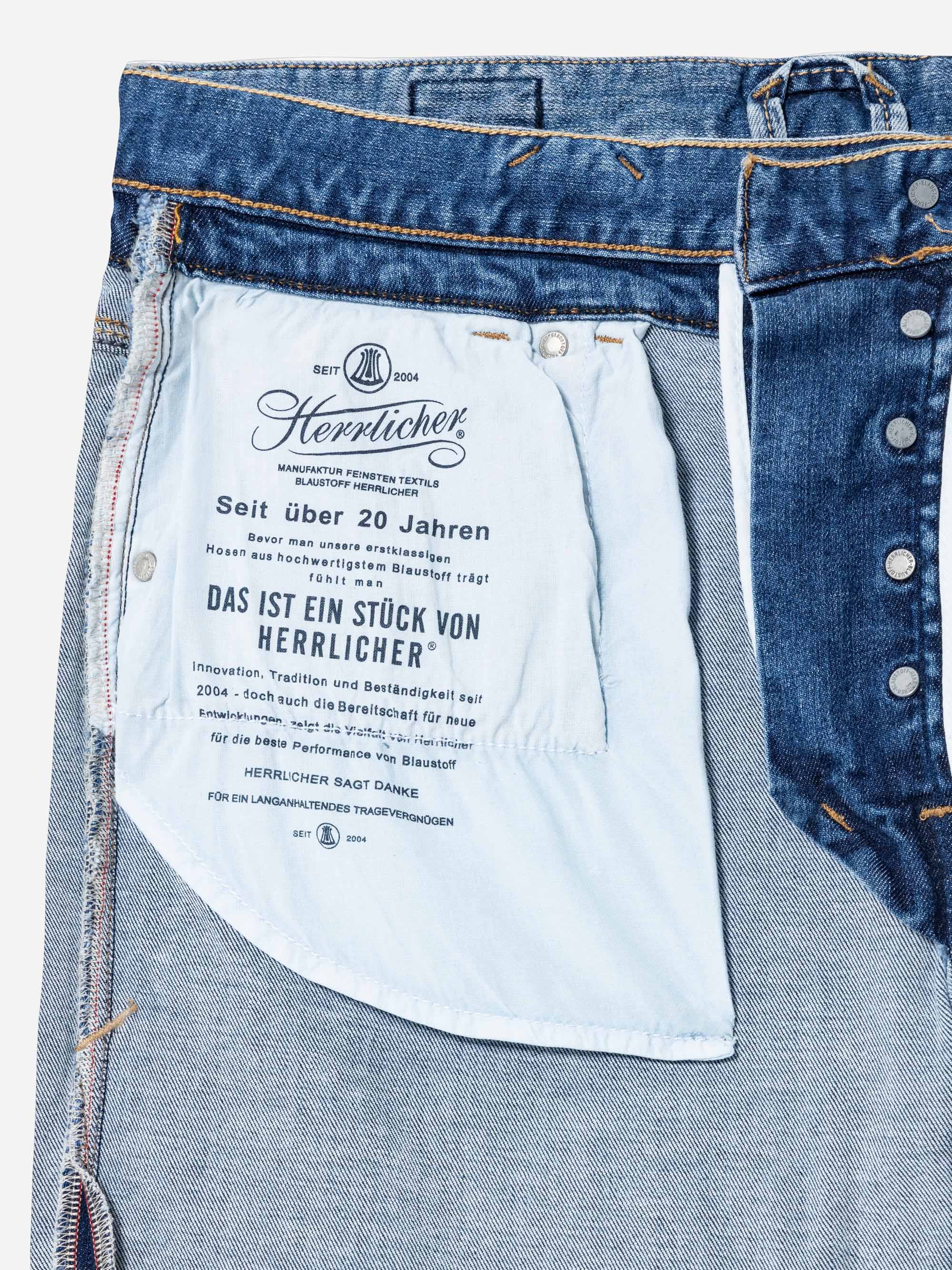 Nahaufnahme der Innenseite einer blauen Jeans, die das weiße Innenfutter der Taschen mit deutschem Text, Metallknöpfen und sichtbaren Nähten zeigt. Im Text werden die Marke Herlicher und Produktinformationen erwähnt.