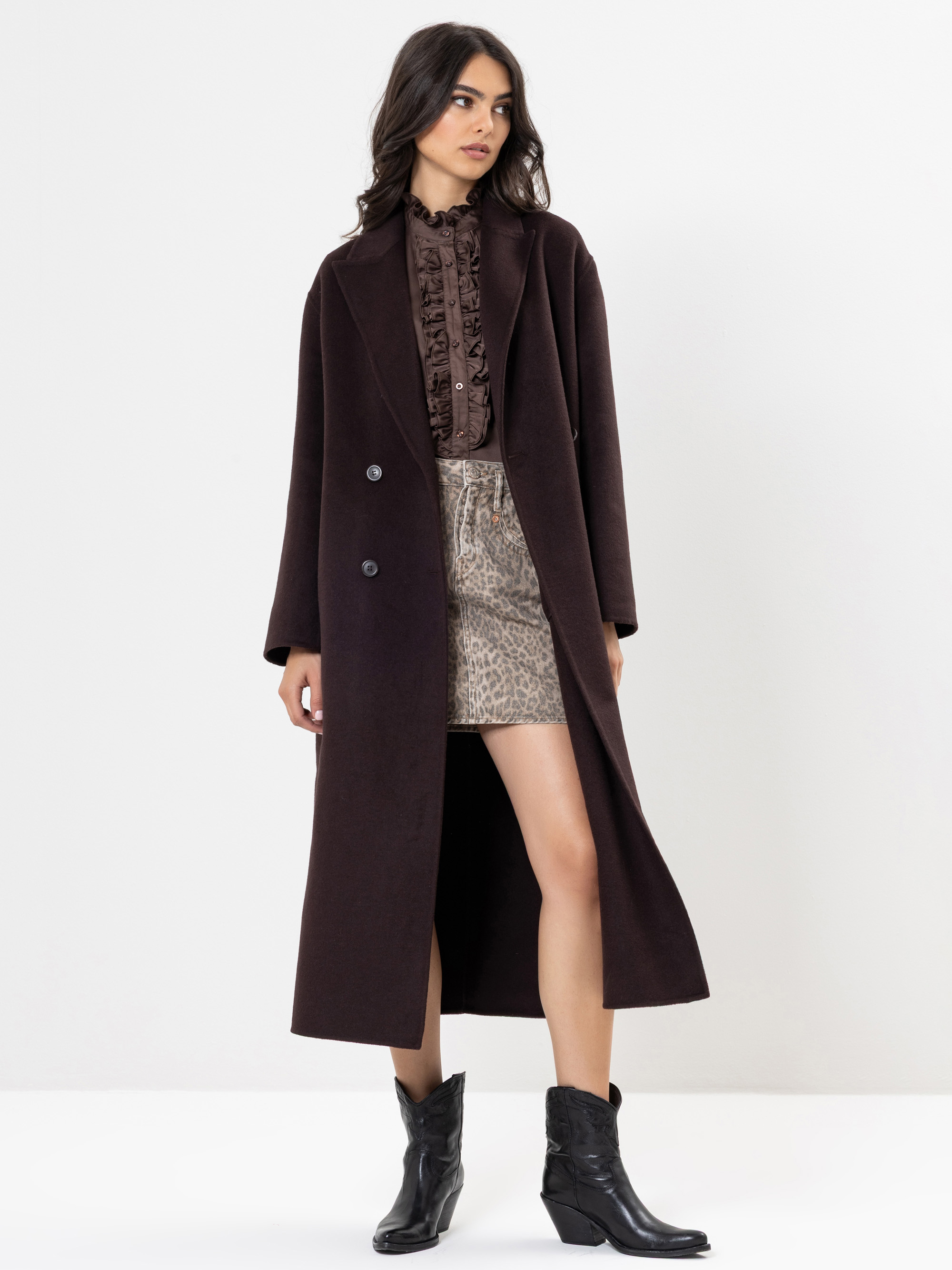 Eine Frau, die einen langen dunkelbraunen Mantel über einer gerüschten braunen Bluse und einen kurzen Rock mit Leopardenmuster trägt, gepaart mit schwarzen Ankle Boots, steht vor einem schlichten weißen Hintergrund.