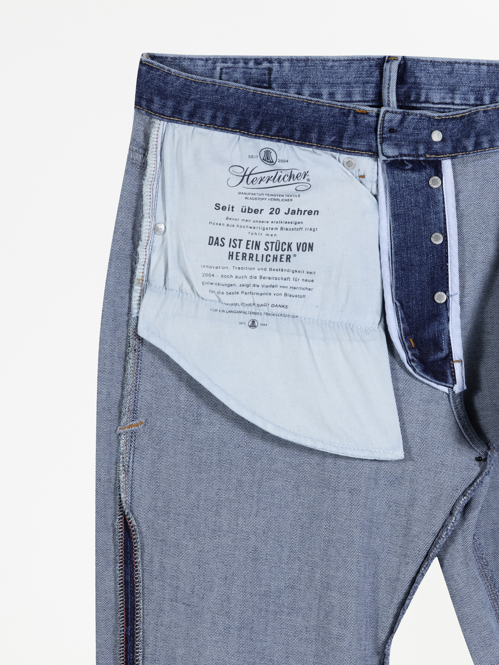 Nahaufnahme der Innenseite einer blauen Jeans, die das Taschenfutter mit aufgedrucktem Text in deutscher Sprache sowie einen Teil des Bunds und des Hosenschlitzes zeigt, alles auf weißem Hintergrund.