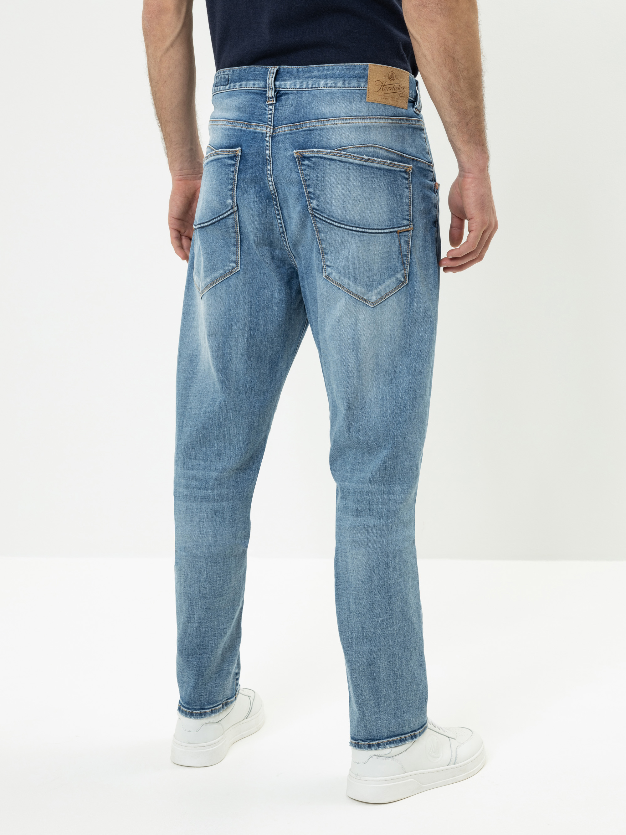 Ein Mann steht mit dem Gesicht zur Wand und trägt eine hellblaue Jeans mit sichtbaren Nähten und Gesäßtaschen, ein dunkles marineblaues Hemd und weiße Turnschuhe vor einem schlichten weißen Hintergrund.
