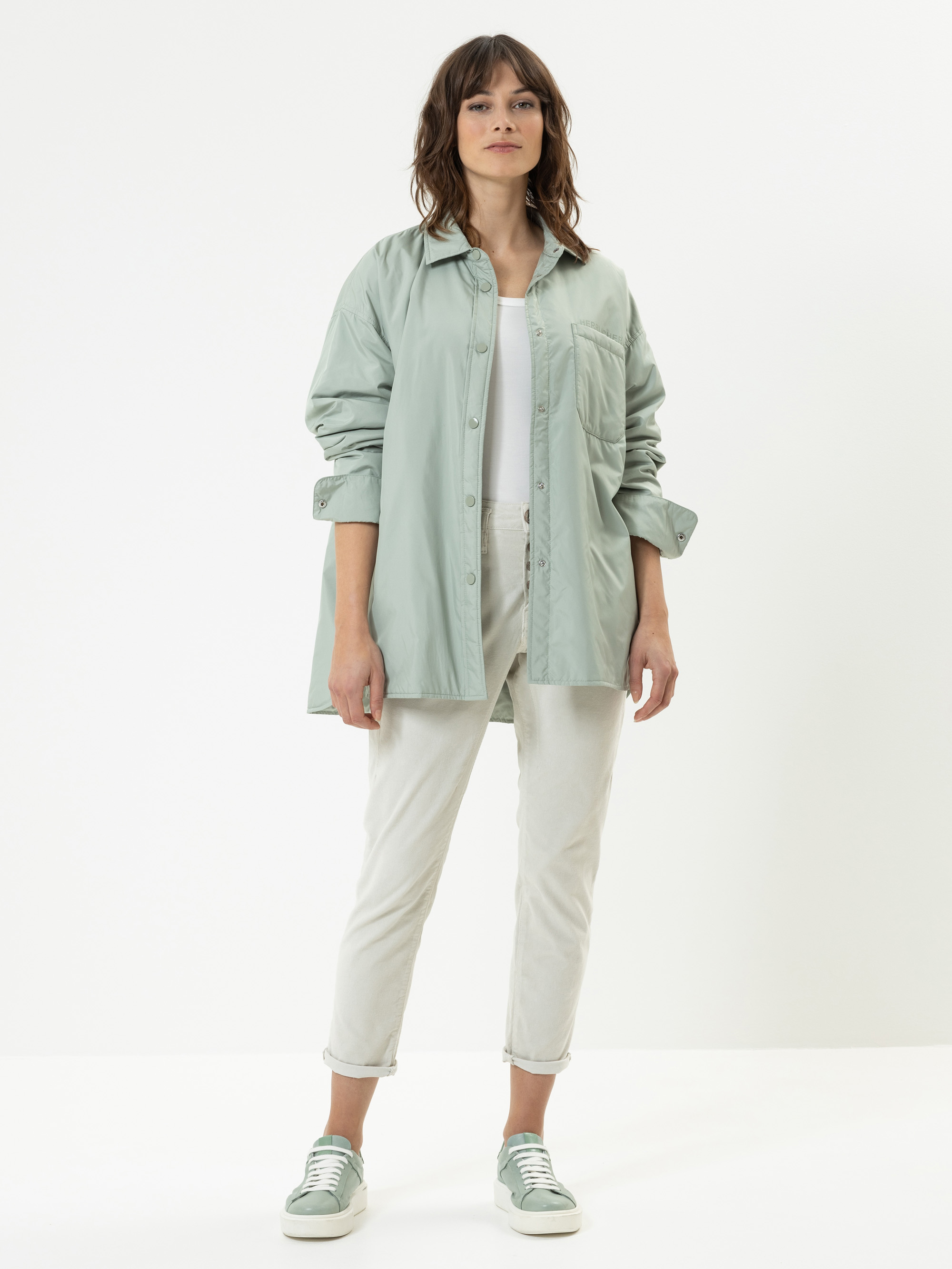 Eine Frau steht mit dem Gesicht nach vorne und trägt ein hellgrünes Button-up-Hemd über einem weißen Oberteil, eine helle Hose und grüne Turnschuhe. Sie hat schulterlanges braunes Haar und einen neutralen Gesichtsausdruck. Der Hintergrund ist einfarb