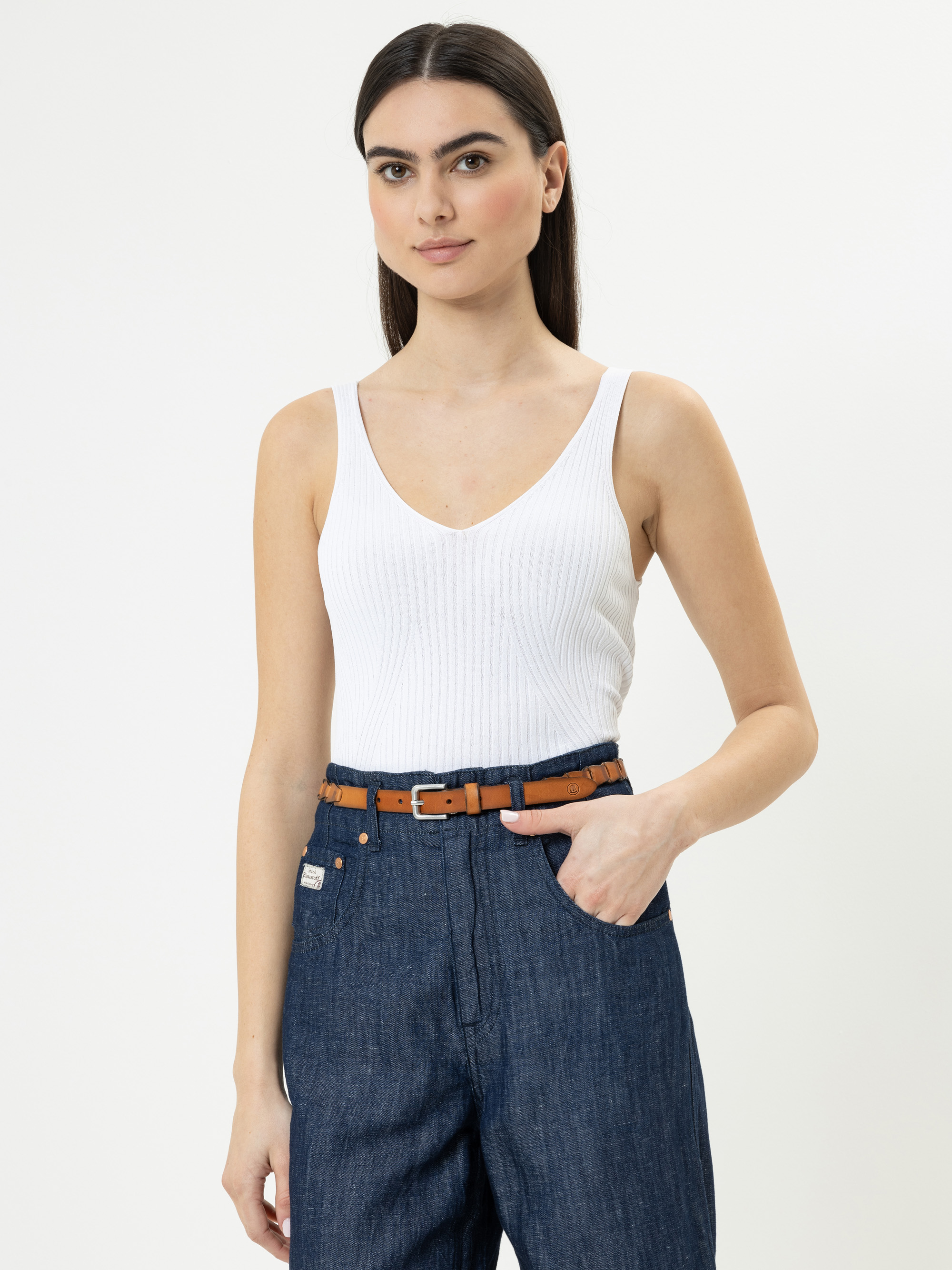 Eine Frau mit langen dunklen Haaren trägt ein weißes, ärmelloses, geripptes Oberteil, das in eine blaue Jeans mit hoher Taille und einem braunen Gürtel gesteckt wird. Sie steht vor einem schlichten weißen Hintergrund und hat eine Hand in der Tasche.