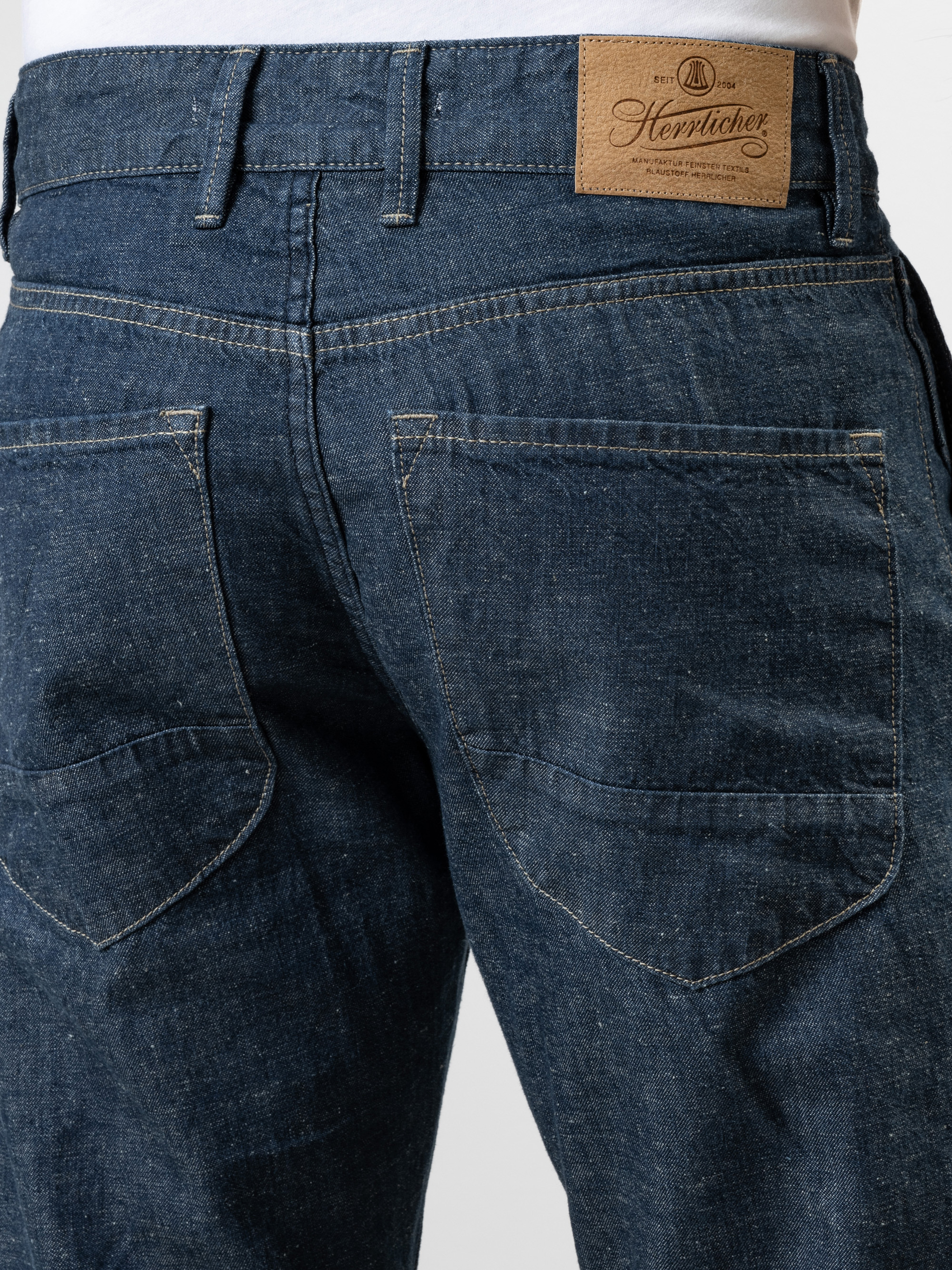 Nahaufnahme der Rückseite einer blauen Denim-Jeans mit zwei Taschen, weißen Kontrastnähten, Gürtelschlaufen und einem braunen Lederaufnäher der Marke Herrlicher am Bund.