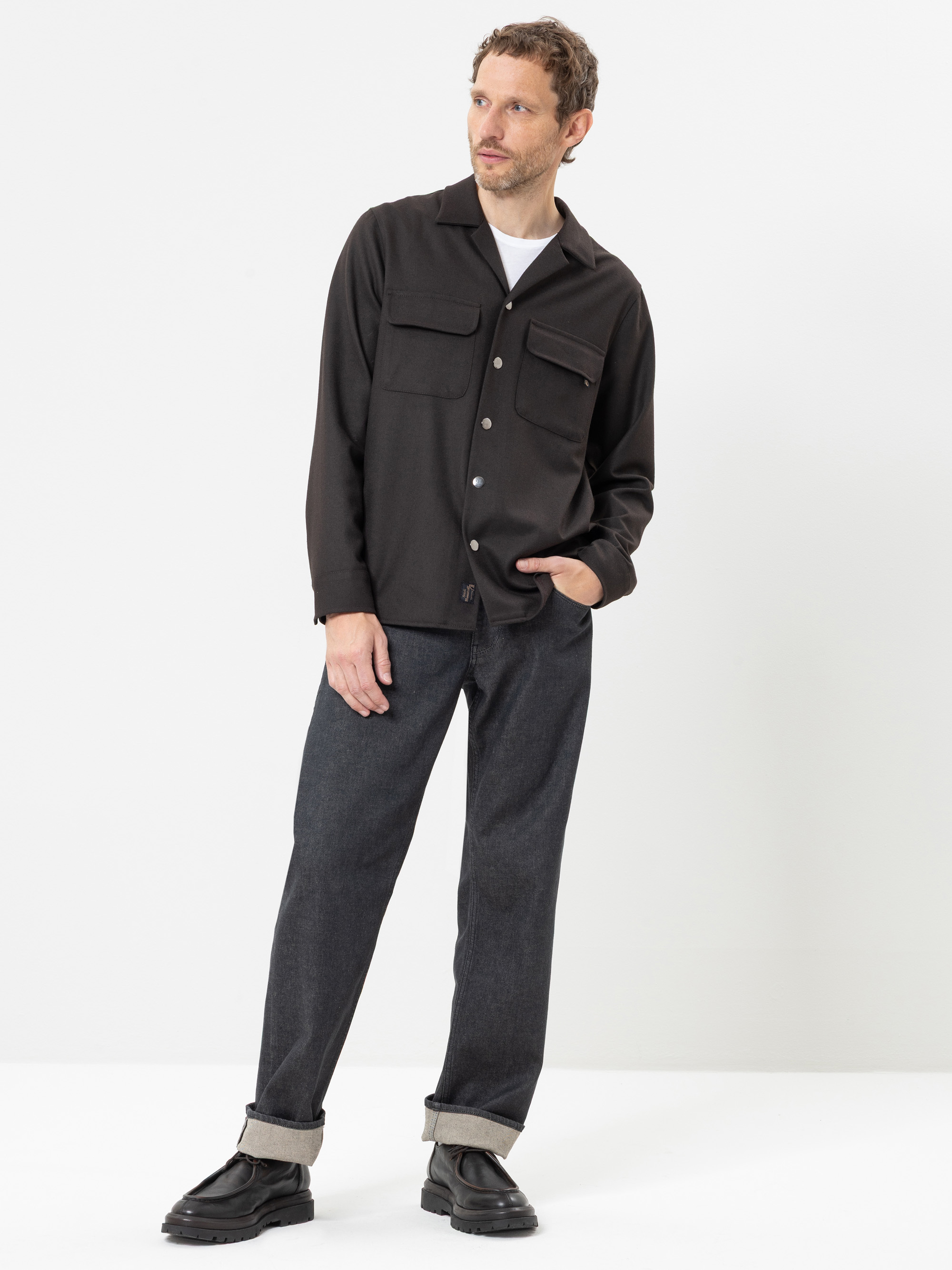 Ein Mann steht lässig vor einem weißen Hintergrund. Er trägt ein schwarzes Hemd mit Knöpfen über einem weißen T-Shirt, eine dunkle Jeans mit Umschlag und schwarze Schuhe. Er hat eine Hand in der Tasche und blickt leicht nach links.