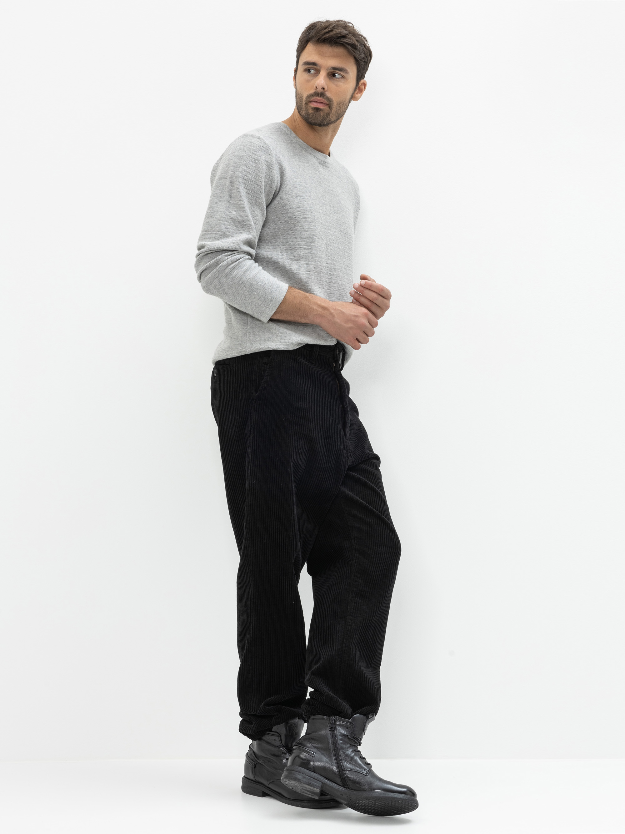 Ein Mann steht vor einem schlichten weißen Hintergrund und trägt ein hellgraues langärmeliges Hemd, eine weite schwarze Hose und schwarze Turnschuhe. Er blickt leicht zur Seite und hat eine Hand vor der Brust erhoben.