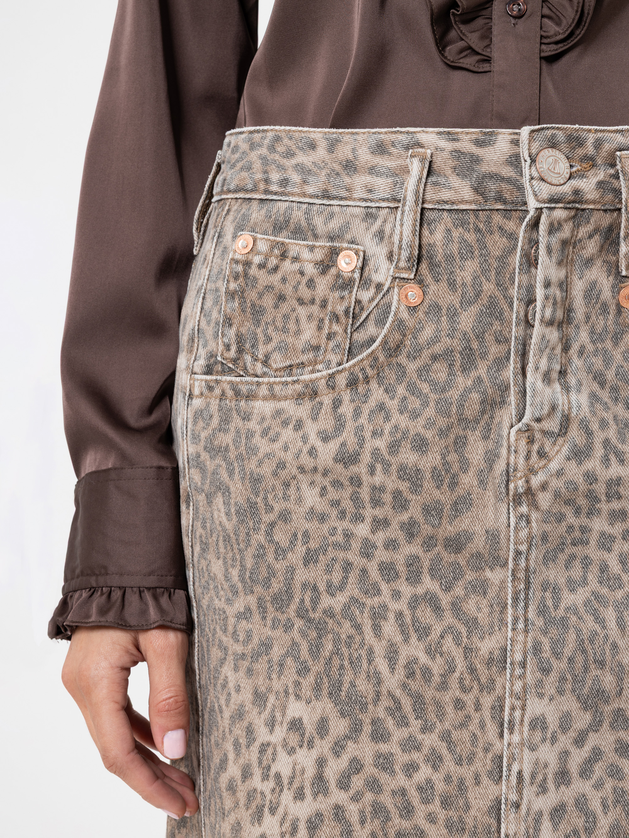 Eine Person, die eine braune, langärmelige Bluse mit gerüschten Manschetten und einen Jeansrock mit hoher Taille und Leopardenmuster trägt. Die Hand der Person ruht an ihrer Seite, und der Hintergrund ist schlicht weiß.