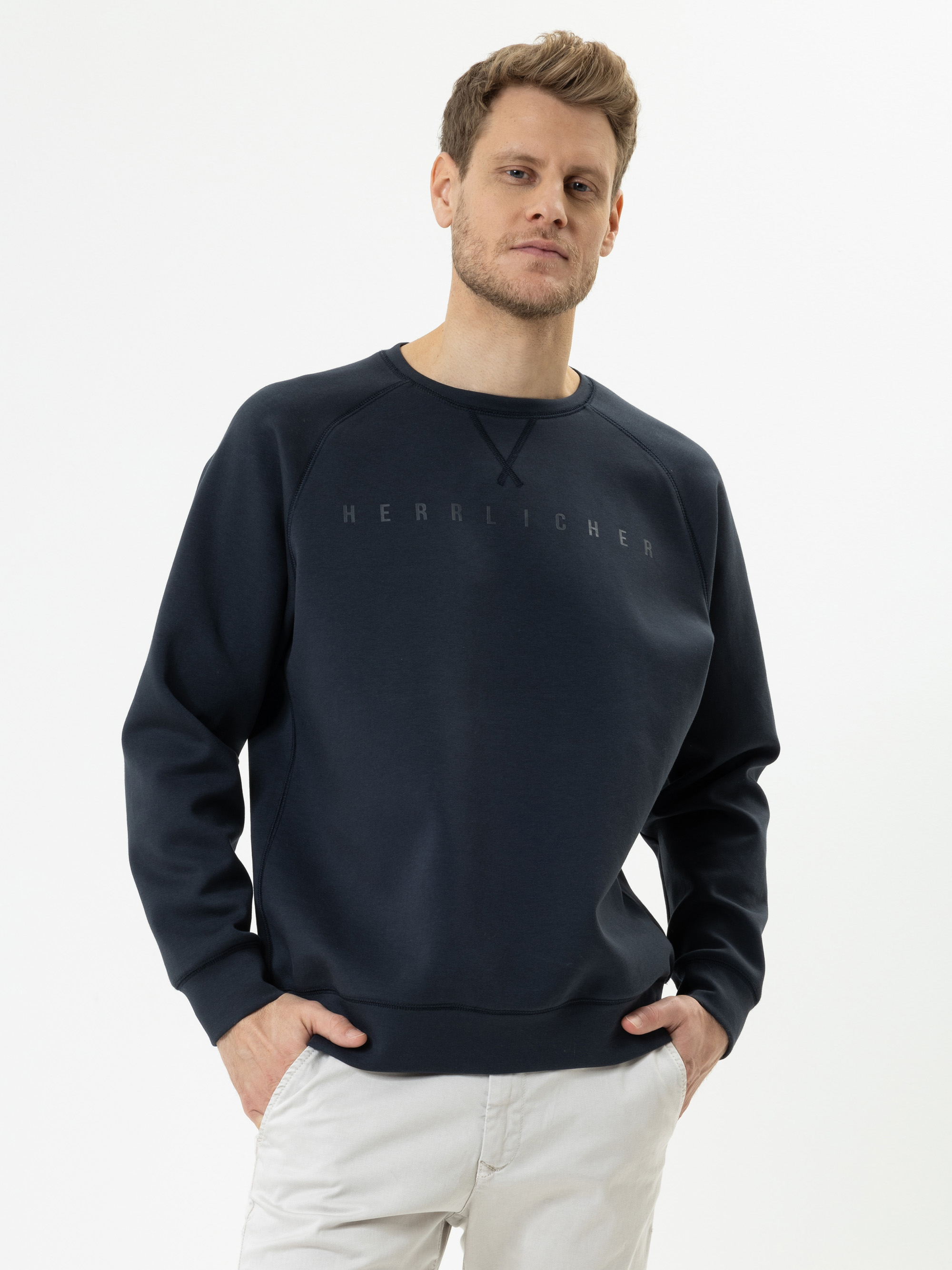Ein Mann in einem dunkelblauen Sweatshirt mit HERRLICHER-Aufdruck auf der Brust steht mit den Händen in den Taschen seiner hellen Hose vor einem schlichten weißen Hintergrund.