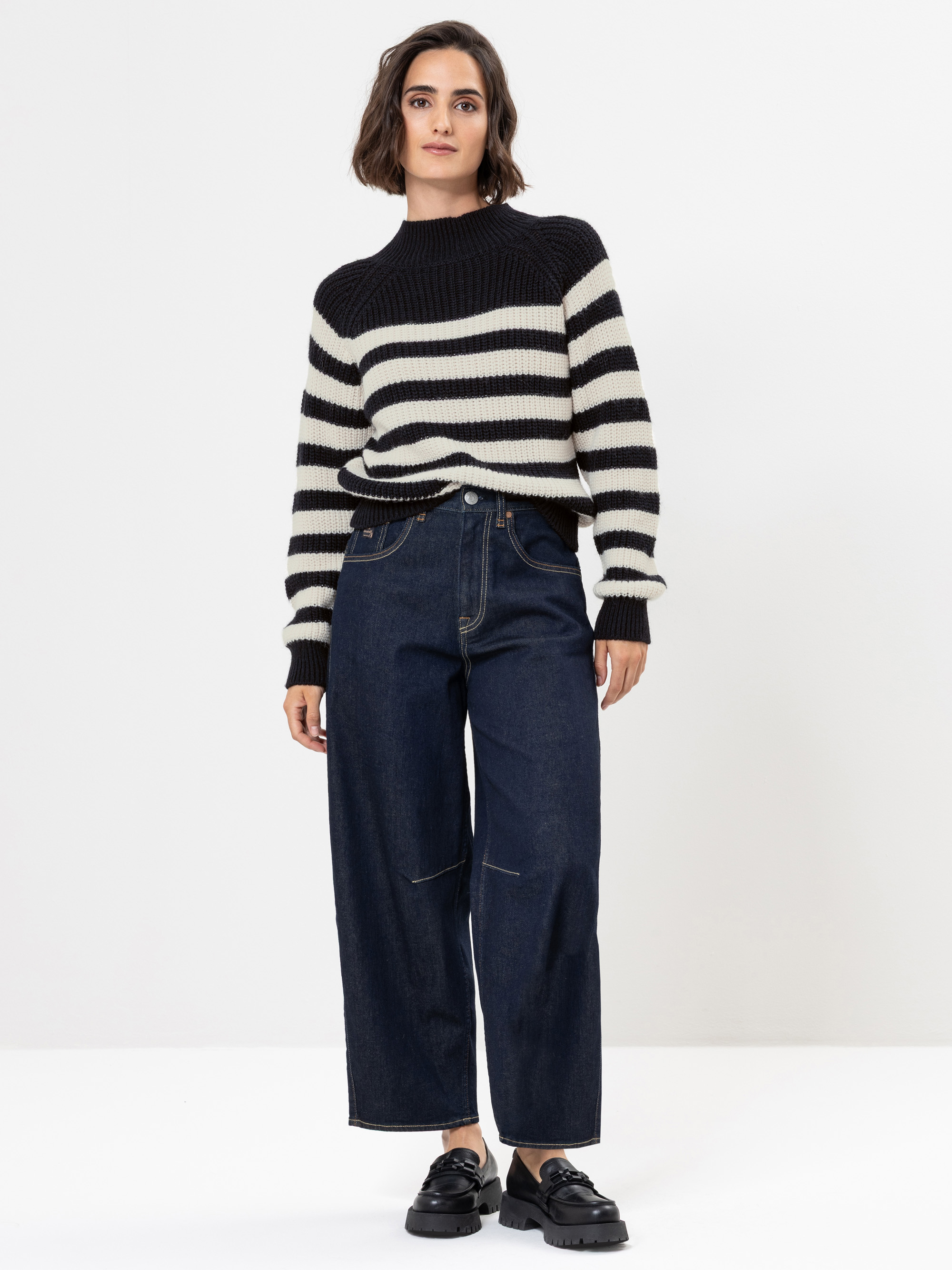 Eine Person steht vor einem schlichten weißen Hintergrund und trägt einen schwarz-weiß gestreiften Pullover, dunkle Jeans mit weitem Bein und schwarze Slipper. Ihr Haar ist schulterlang und gewellt.