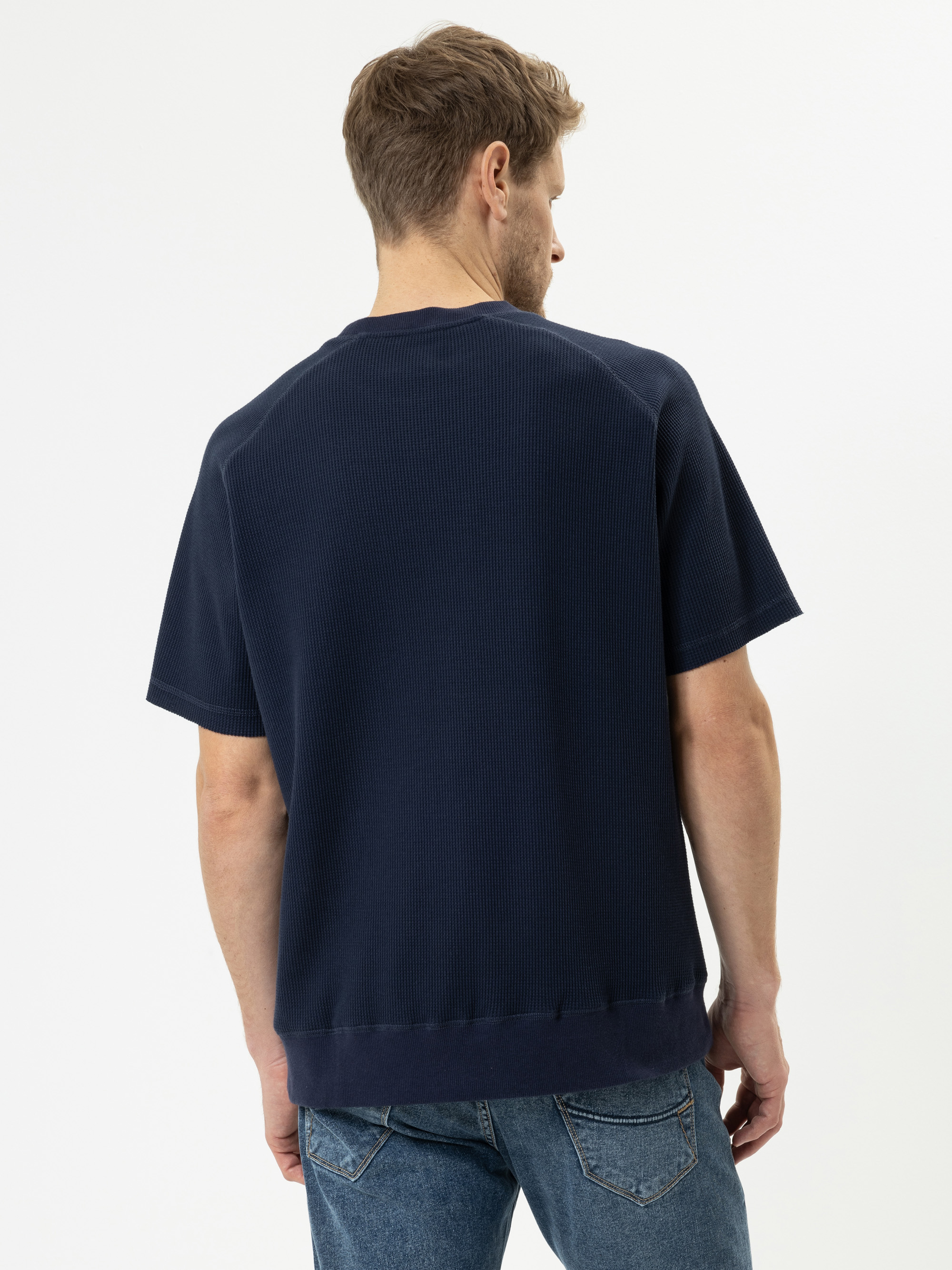 Ein Mann mit hellbraunem Haar ist von hinten zu sehen. Er trägt ein marineblaues, kurzärmeliges Hemd und blaue Jeans vor einem schlichten weißen Hintergrund.